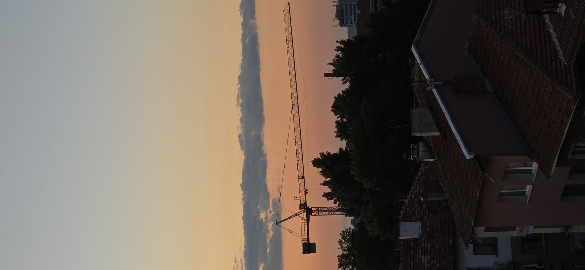 In the city of Unknown, Unknown, a construction crane stands silhouetted against a gradient sky transitioning from pale blue at the top to warm orange and pink hues near the horizon. A line of dark clouds stretches horizontally across the sky just above the crane's jib. Below the crane, a dense canopy of dark green trees obscures the lower parts of the cityscape. In the foreground, the tiled roofs of residential buildings are visible, some with chimneys. The lighting suggests it is either sunrise or sunset. There are no people clearly discernible in the image.
