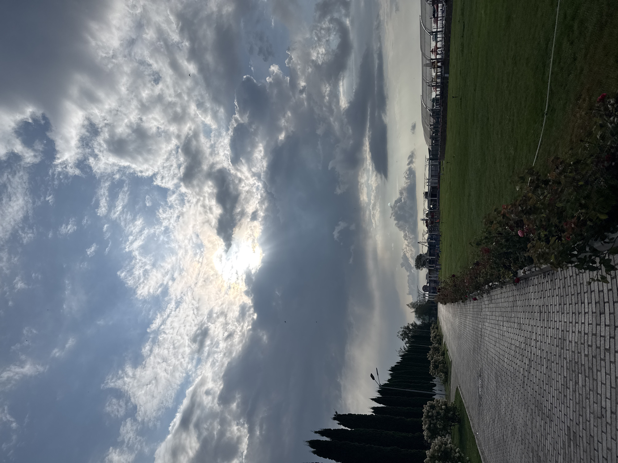 The image depicts a paved walkway in the city of Sary-Oy, Kyrgyzstan, flanked by a manicured lawn on the right and a row of manicured bushes and tall, dark green trees on the left. The walkway curves to the right as it recedes into the distance. In the background, there are low-lying buildings with arched roofs, some structures resembling playground equipment, and a large expanse of green grass. The sky above is dramatic, with a mix of dark, heavy clouds and brighter, scattered clouds, through which the sun is breaking, casting rays of light. A few dark specks, likely birds, are visible in the sky. The overall impression is of an outdoor recreational or park area under a dynamic sky.