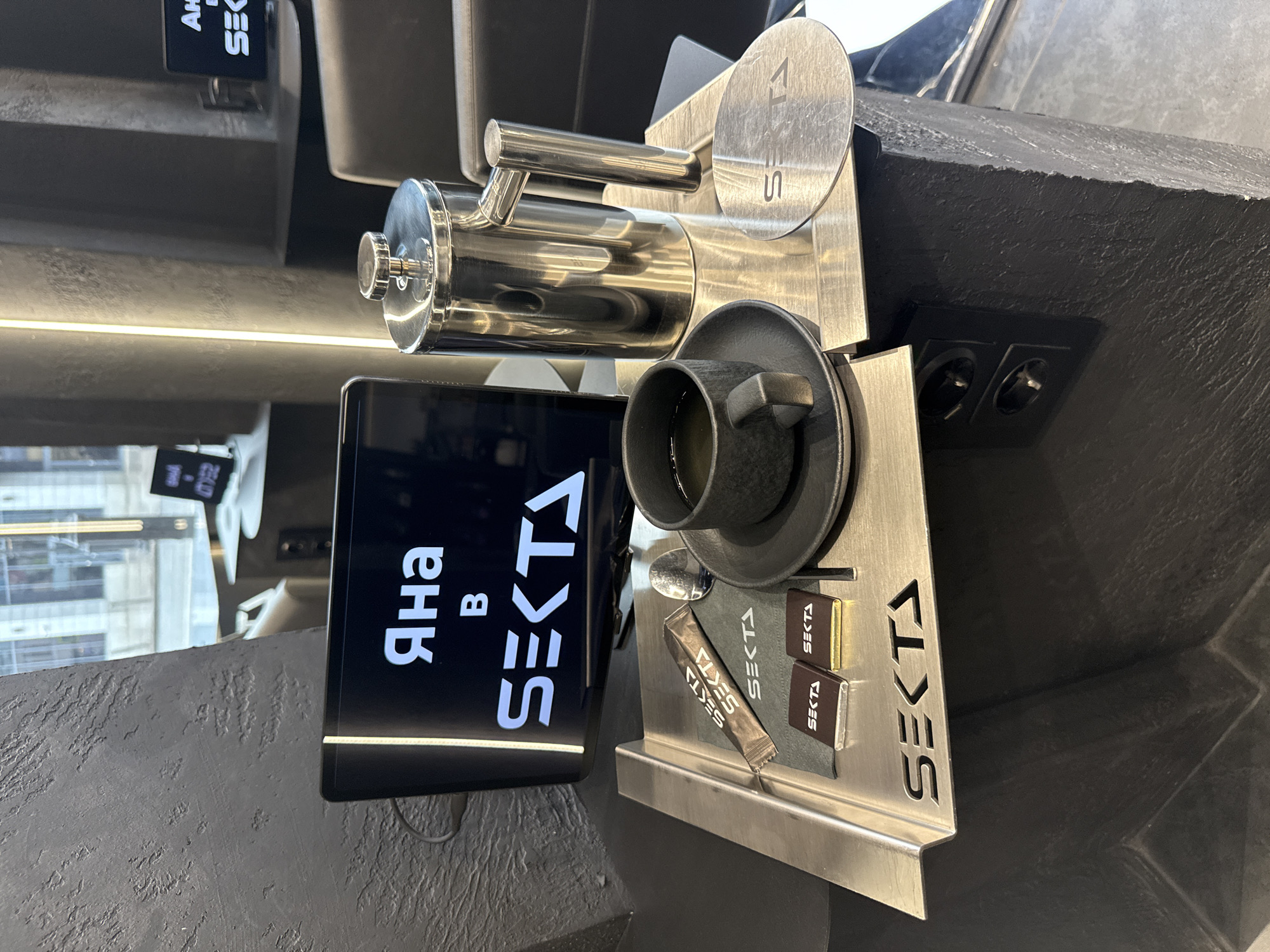 The image shows a coffee setup with branding for "SEKTA", located in what appears to be a cafe. There is a metal espresso machine, a black coffee cup on a saucer, a digital display showing the name "SEKTA" in Russian, and sugar or sweetener packets. There are electrical outlets in the background. The setup is on a textured, dark gray surface. Through the window, a glimpse of a cityscape is visible. Based on the branding and architectural context, the location is likely Moscow, Russia.