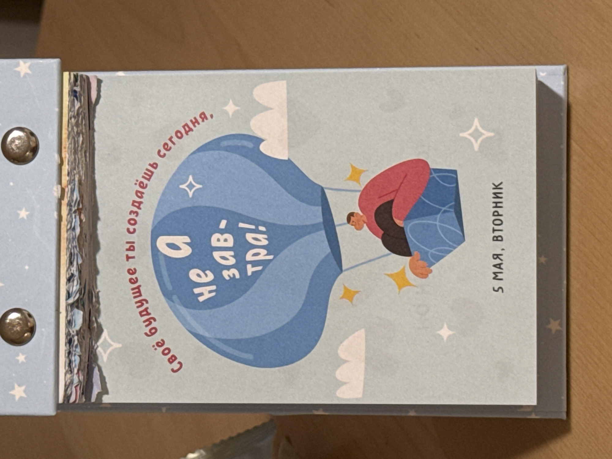 The image shows a page from a desk calendar. The illustration features a person in a red shirt inside a blue hot air balloon with the text "Своё будущее ты создаёшь сегодня, a не завтра!" which translates to "You create your future today, not tomorrow!" The calendar indicates the date "5 мая, вторник" which is May 5, Tuesday. The calendar is on a table, likely indoors. There is no indication of the location being Izhevsk, Russia or of any additional people, subjects, or actions.