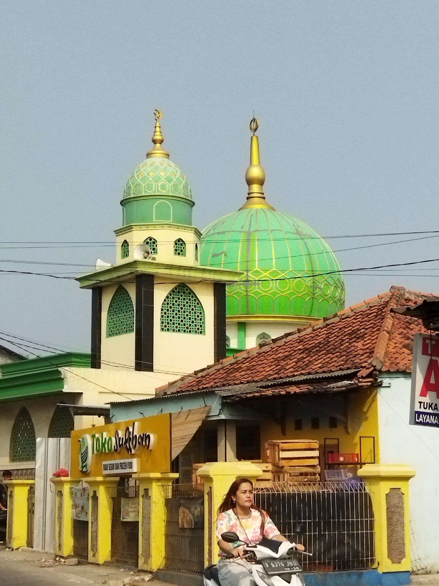 In the city of Suka Sari, Indonesia, a mosque with green domes and gold accents stands prominently against a clear sky. In the foreground, a woman rides a motorcycle past a yellow fence and a building with a sign that reads "Toko Suliban." The scene depicts a typical street view in a residential or commercial area. The mosque's architecture, with its minaret and decorative domes, suggests a place of worship. The presence of a local shop and the woman on the motorcycle indicate daily life in the community. The overall image captures a slice of Indonesian urban landscape, blending religious structures with everyday activities.