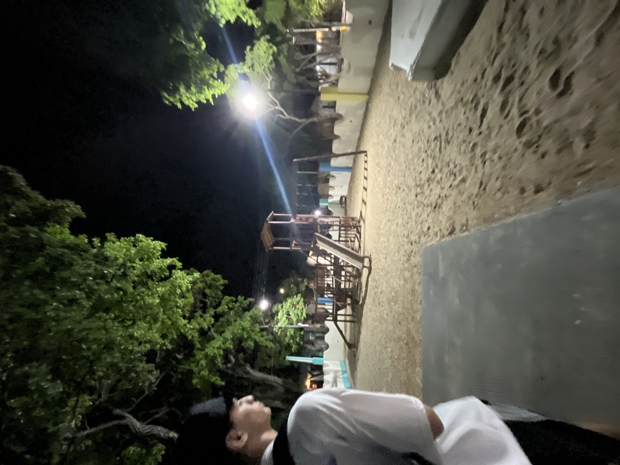 An outdoor playground at night in Santa Ana, Venezuela is the subject of this image. A person wearing a white t-shirt and a black baseball cap stands in the foreground, looking to the left of the frame. The playground features a wooden play structure with slides and swings, surrounded by a sandy ground. A low, decorative wall painted with white, blue, and yellow sections lines the perimeter of the playground. Trees with lush green foliage are visible in the background, with lights illuminating the scene and casting shadows. The overall atmosphere suggests a quiet and calm evening.