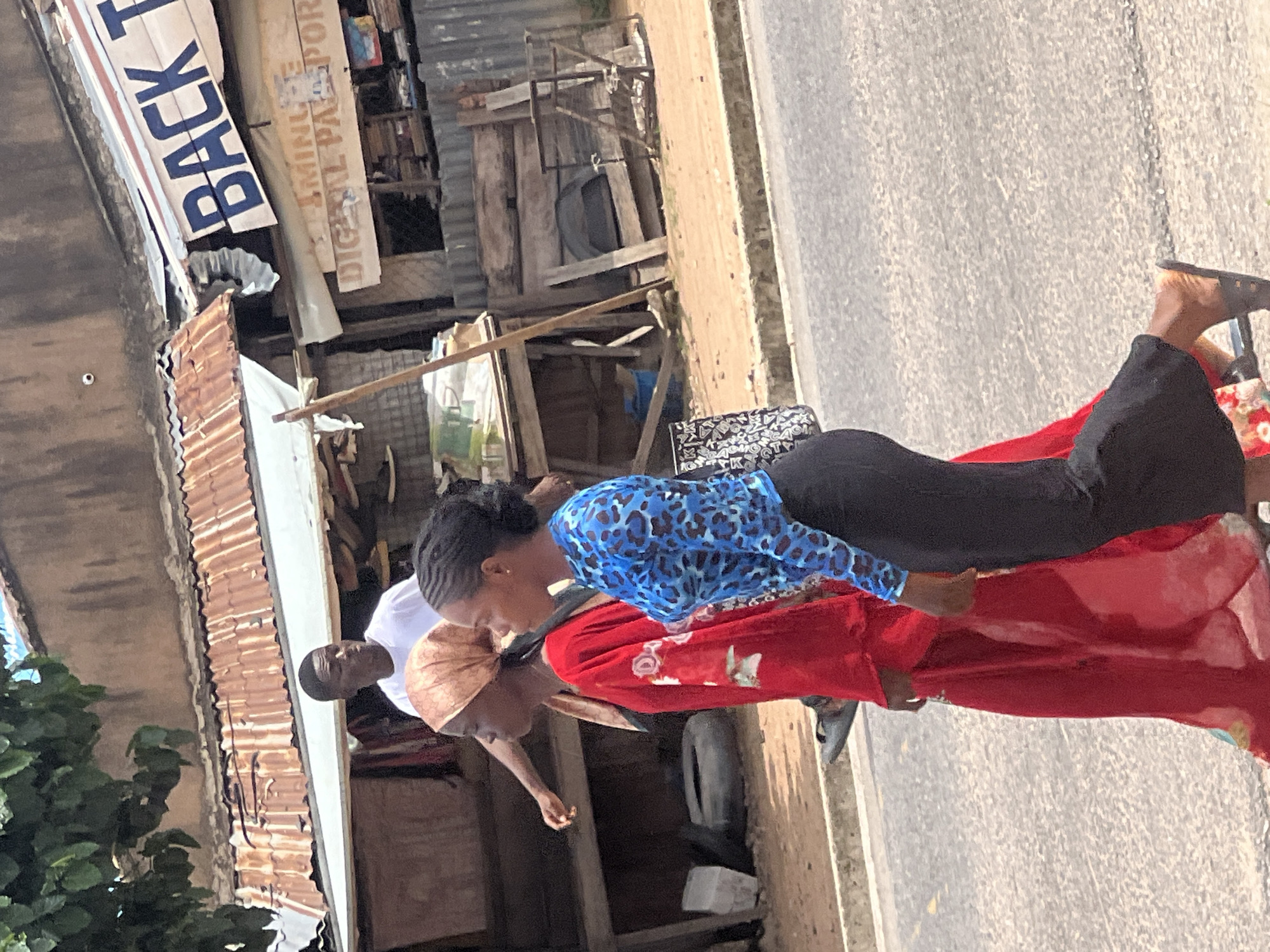 In Jalingo, Nigeria, three individuals are captured in a street scene. A Black male, wearing a white t-shirt, walks behind two Black females. The female closer to the viewer is wearing a blue, leopard-print long-sleeved shirt and black pants, carrying a black and white patterned bag. The second female, walking to her left, is adorned in a red, flowing dress and a tan headscarf. They are walking on a paved surface adjacent to a roadside with shops. The shops have corrugated metal roofs and wooden structures. One shop sign reads "BACK T". Another sign is partially visible and appears to include the words "DIGITAL PASPOR". The environment suggests a commercial area with modest infrastructure. The lighting indicates it is daytime, with sunlight casting shadows.