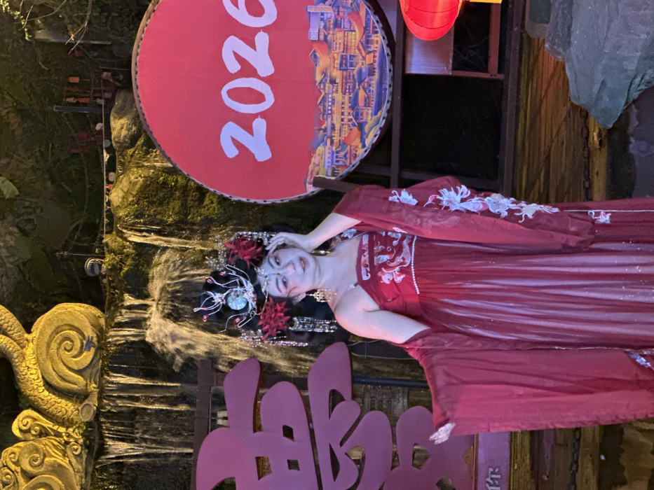 A female individual is depicted in a full-length, slightly off-center vertical shot at an outdoor night scene in Yuzhong District, China. The subject, appearing to be of East Asian descent, wears an elaborate traditional Chinese-style outfit, primarily red. This attire consists of a strapless inner garment with white and light blue embroidery on the bodice, a shimmering floor-length red skirt, and a matching long-sleeved outer robe with similar white and light blue embroidery on the sleeves. Her head is adorned with a complex silver and red headpiece featuring a crown-like structure, red floral accents, and numerous dangling ornaments. She also wears long, ornate gold-colored earrings. Her left hand is raised to her left temple, fingers slightly curled, while her right arm is extended outwards with an open hand. Her expression is a subtle smile directed at the camera.
The background features an illuminated artificial waterfall cascading over dark rock formations, above which a golden, coiled, serpentine sculpture is visible on the upper left. To the subject's left, a large, illuminated pink-red Chinese character is partially visible. To her right, a prominent circular red sign displays the white numerals "2026" above an illustration of traditional Chinese architecture. This sign is mounted on a dark wooden frame. A red, traditional Chinese lantern is partially visible on the far right foreground. The environment is illuminated by artificial light sources.