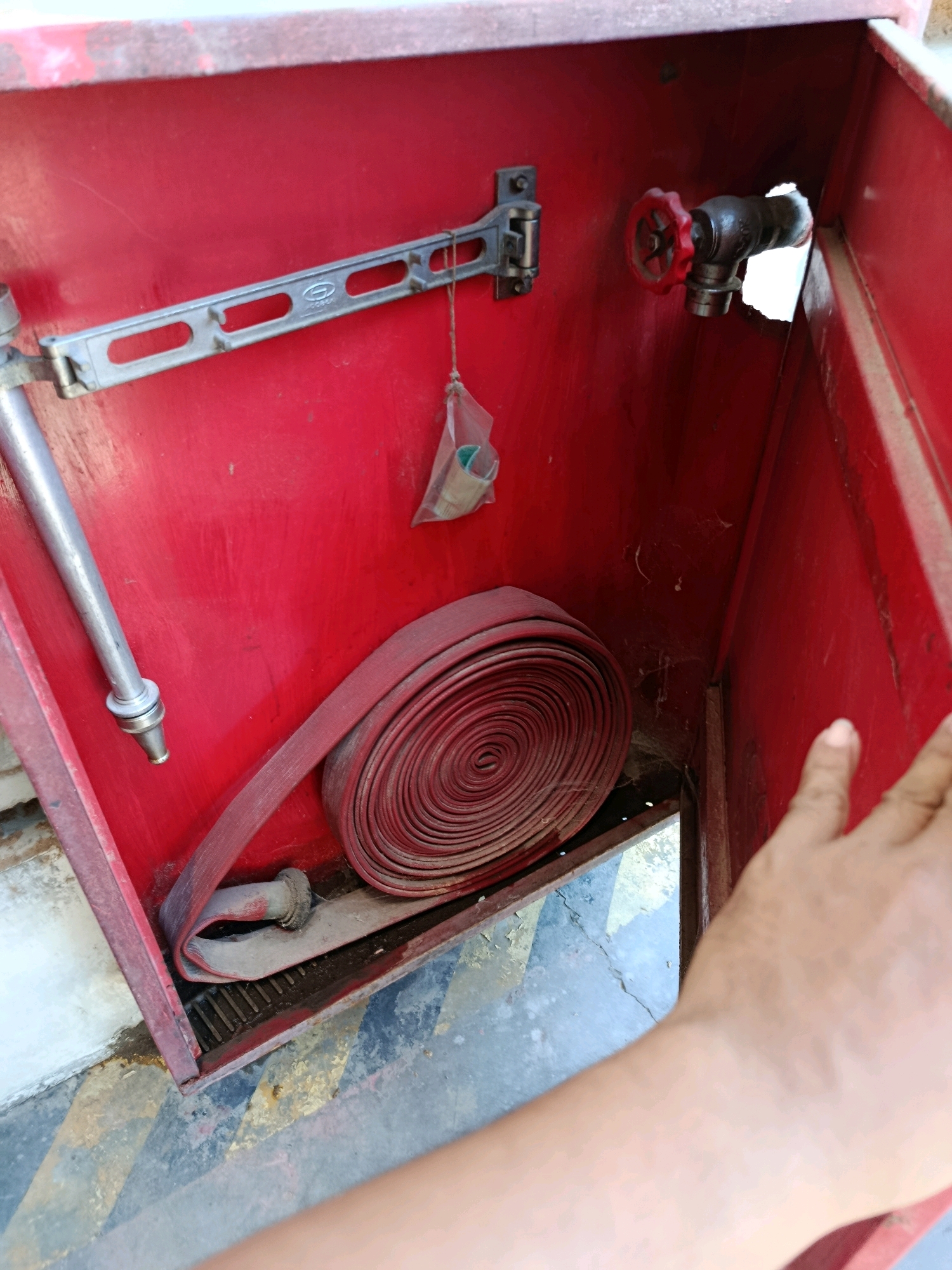 A first-person perspective shot shows a hand reaching into a red fire hose cabinet. The cabinet is open, revealing a coiled red fire hose resting on a slatted metal base. To the left, a silver metal hose nozzle is attached to a pipe. On the back wall of the cabinet, a silver bracket with oblong holes is mounted, and a small plastic bag containing what appears to be a green object hangs from it by a string. On the right side of the cabinet's back wall, a black pipe with a red-handled valve is visible, protruding from a hole in the wall. The floor beneath the cabinet has a patterned surface of black and yellow diagonal stripes. The overall environment suggests an outdoor urban setting, potentially within the city of Kembangarum, Indonesia, given the context.