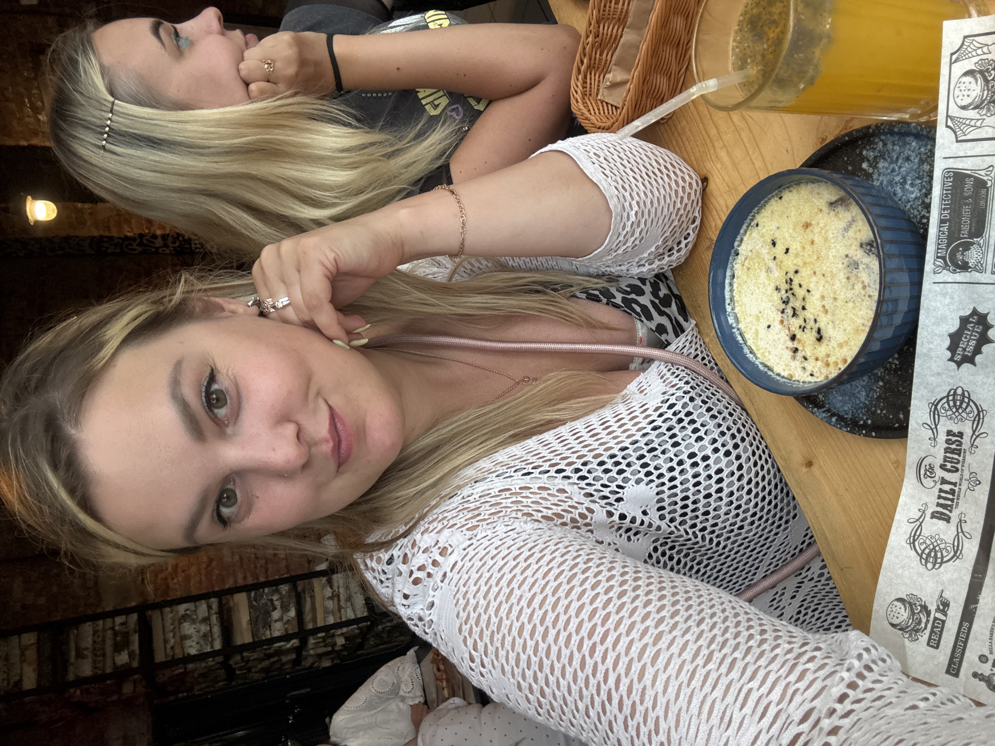 Two women are pictured in a selfie. The woman in the foreground is blonde and wearing a white, crocheted top over a black leopard print garment. She has blonde hair and is looking at the camera, smiling. The woman in the background is also blonde and wearing a black t-shirt. She is looking to the side and resting her head on her hand, appearing contemplative.
They are seated at a wooden table, likely in a restaurant or cafe. On the table, a bowl of soup or a creamy dish with black seeds is visible, along with a glass of orange juice and a woven basket. A folded newspaper or menu, with the text "The DAILY CURSE" and "MAGICAL DETECTIVES" prominently displayed, is also on the table. The background shows hints of a brick wall and possibly some metal railings, suggesting an indoor, possibly rustic or industrial, setting. The lighting appears to be artificial, possibly from overhead lamps, indicating an indoor environment at any time of day. The women's relaxed postures and smiles suggest a casual outing and a positive mood.