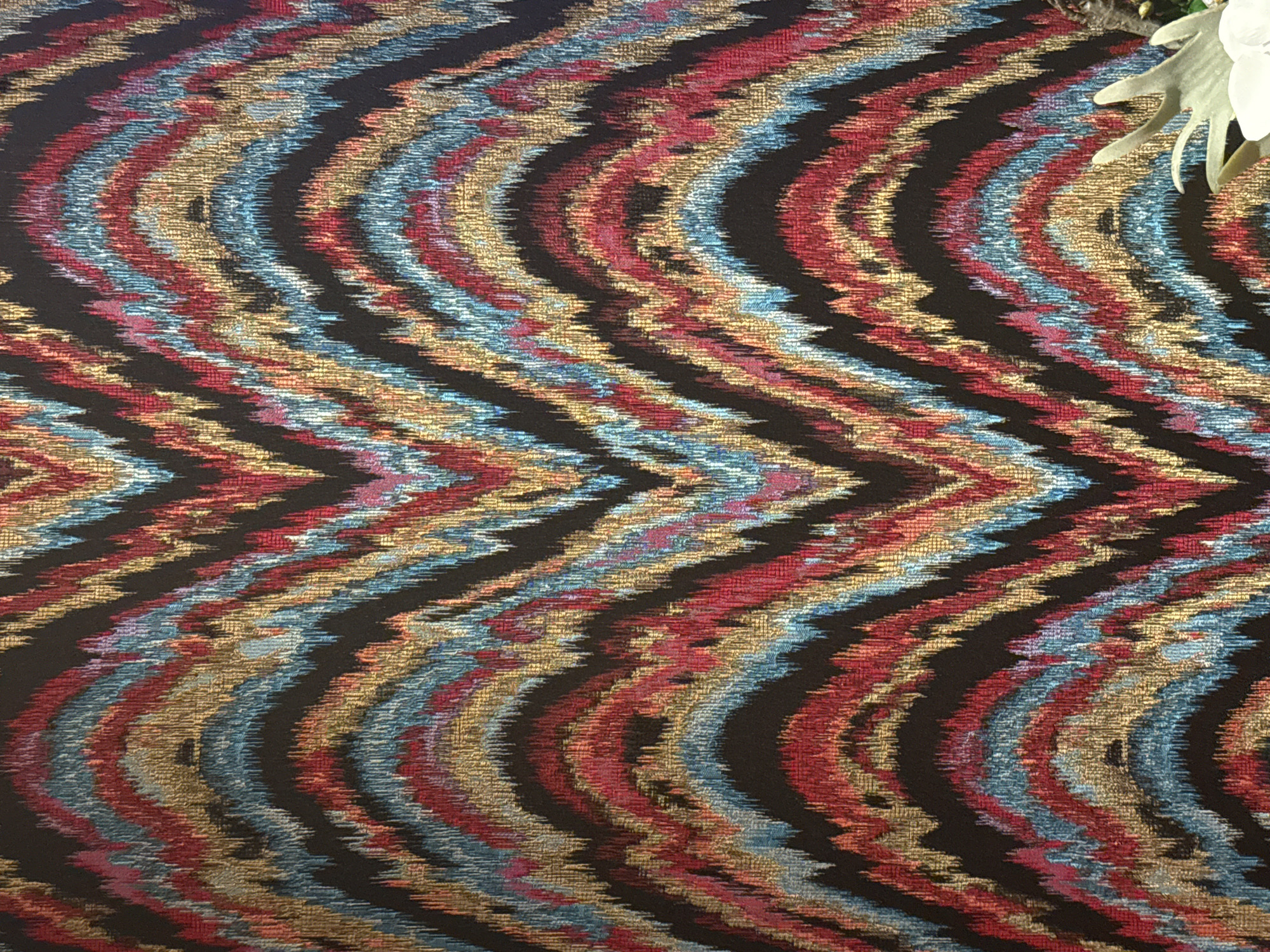 The image is a close-up, overhead shot of a textured fabric with a wavy, zigzag pattern. The fabric features bands of black, gold, red, and blue threads, creating a vibrant, somewhat abstract design. In the upper right corner, a portion of a white flower and some grey, twig-like elements are visible, suggesting the fabric might be part of a decorative arrangement or a piece of clothing with floral embellishments. The lighting is consistent across the fabric, indicating an indoor setting with diffused light. No people, specific activities, text, or discernible location cues beyond the material itself are present in the image.