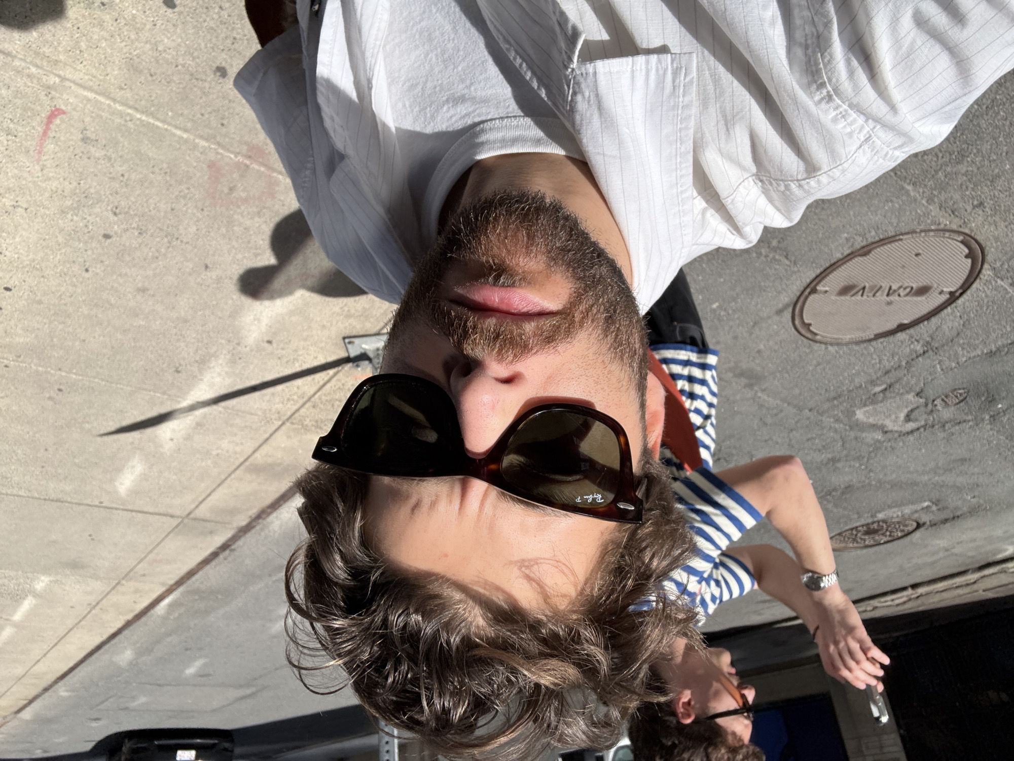 A man is on a sidewalk in Boston, United States of America. He has dark hair and a beard, and he wears sunglasses and a white shirt. Another man is in the lower right, wearing sunglasses and a striped shirt. A manhole cover is in the upper right corner.