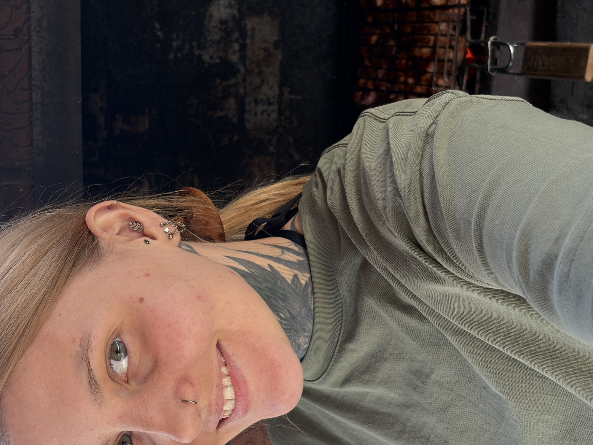 A selfie shows a blonde woman with multiple ear piercings and a nose ring. She is wearing an olive-green shirt. Her neck is tattooed with a black design. In the background, a grill with meat is visible. The location is Chaikovsky, Russia.