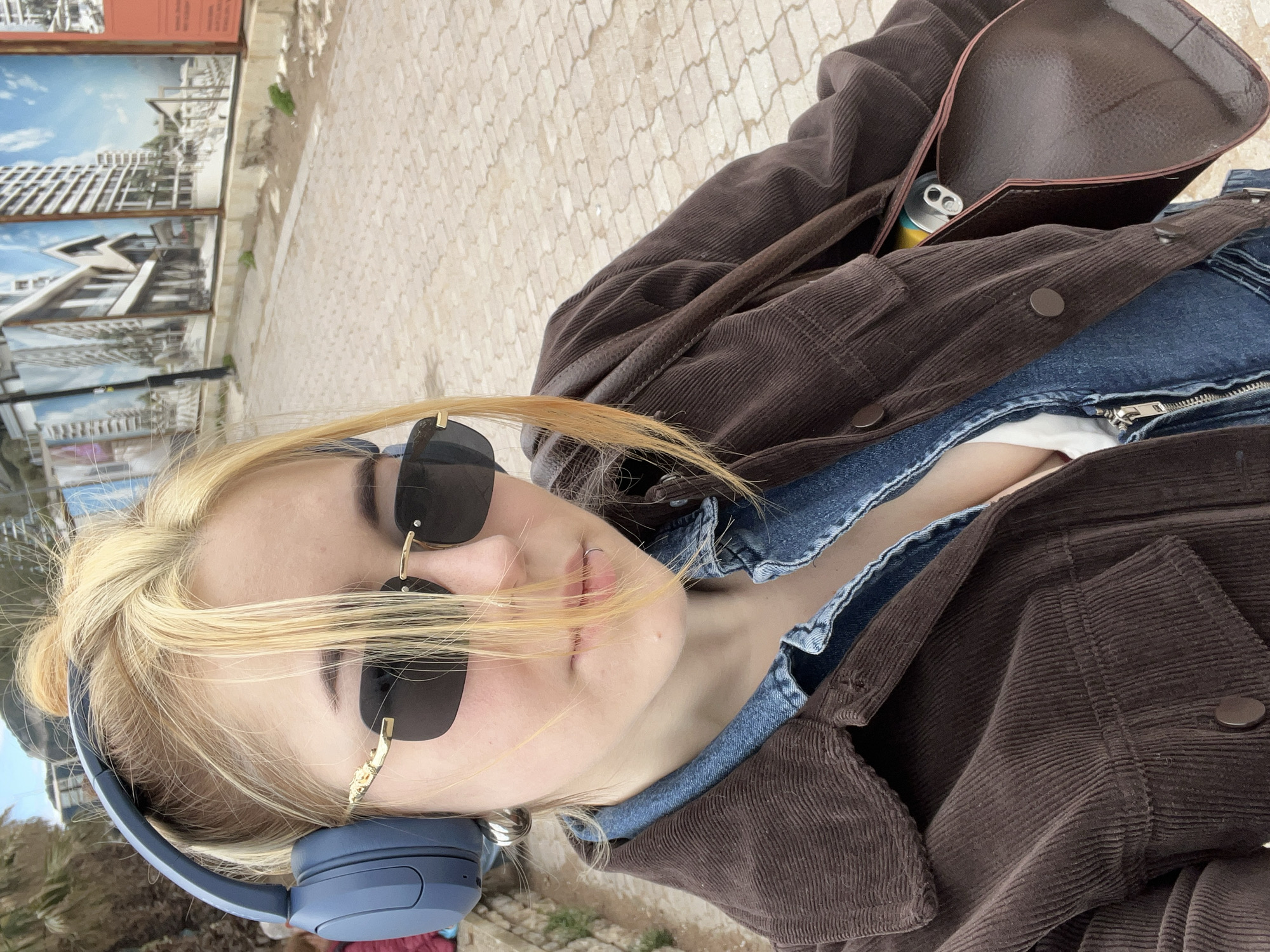 A young woman stands outdoors on a light-colored, patterned stone surface. She has blond hair and wears dark sunglasses and blue headphones. She is also wearing a brown corduroy jacket, a blue denim shirt, and a white t-shirt. A brown leather bag with a can inside hangs off of her shoulder. In the background are framed architectural renderings, suggesting this could be a display area or construction site in Rafailovići, Montenegro.