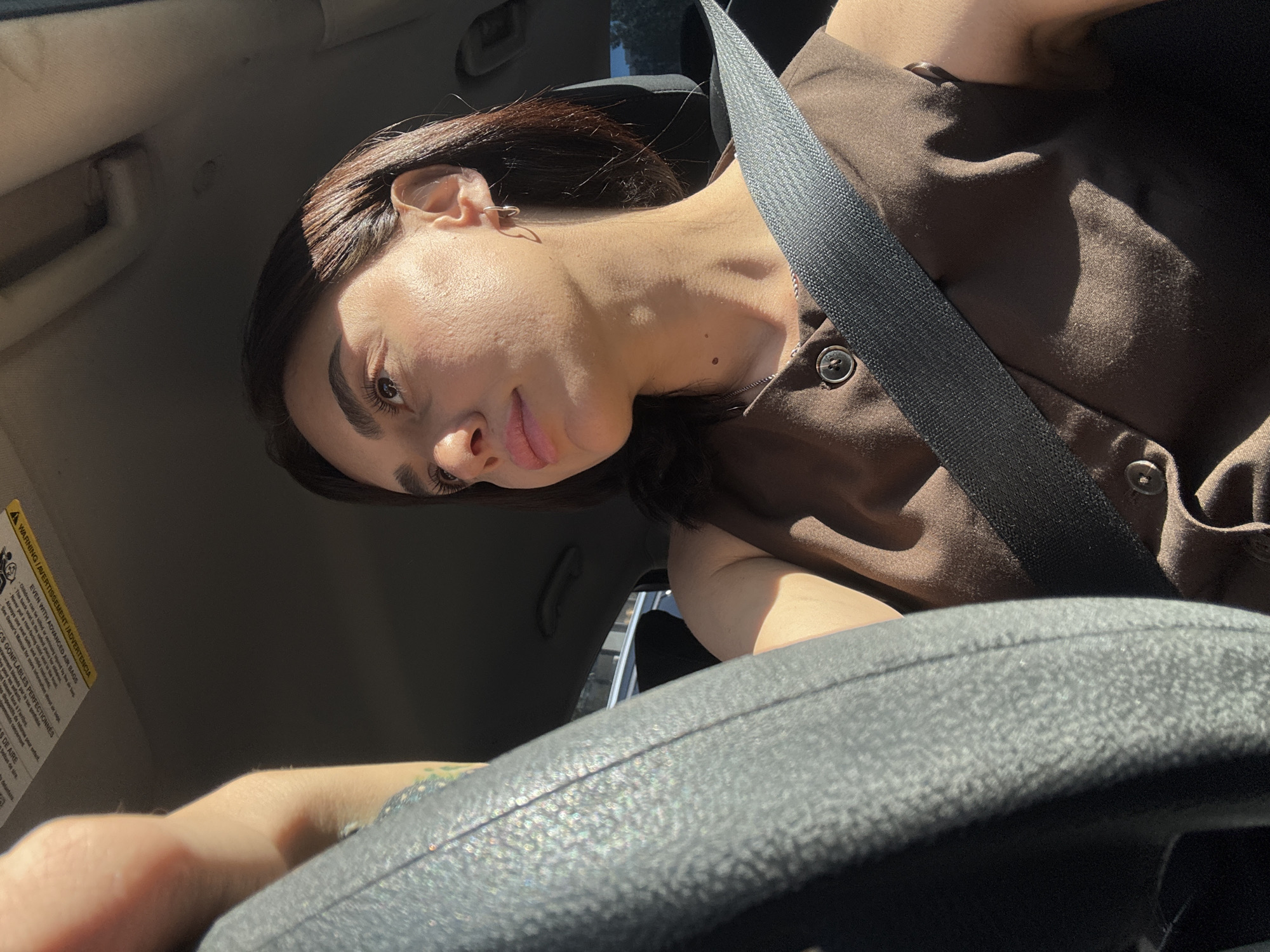 A woman is captured in a close-up shot, seated in the driver's seat of a car, with her hand on the steering wheel. The interior of the car, including the dashboard and a portion of the ceiling with warning labels, is visible. The woman's face is turned away from the camera, looking to the right, with sunlight illuminating her features. She is wearing a brown sleeveless top with two buttons and a seatbelt across her chest. The background outside the car is blurred, showing a hint of buildings and trees, suggestive of an urban or suburban environment. The overall lighting indicates it is daytime. No specific city landmarks or details allowing for definitive identification of Tbilisi, Georgia are present in the image.