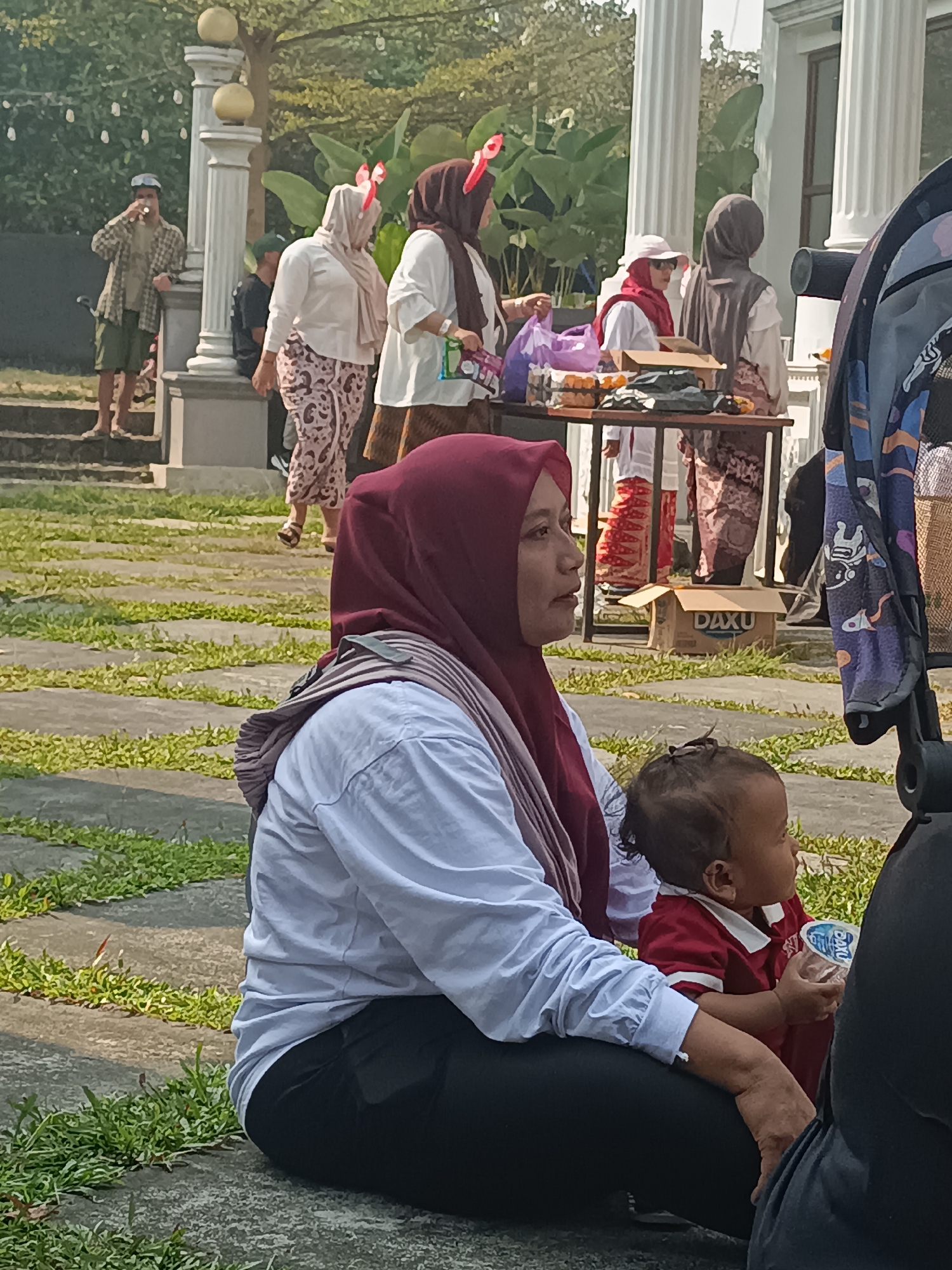 In Sleman, Indonesia, a woman wearing a maroon hijab and a light blue long-sleeved shirt sits on the ground, holding a child wearing a red shirt. The child is holding a plastic cup. In the background, several other people, predominantly women in hijabs, are standing near a table laden with various items, possibly food or goods, suggesting an outdoor gathering or market. One person is smoking and standing on stairs. The environment appears to be a park or an outdoor event space, with stone pathways and grass. A stroller is partially visible to the right of the frame.