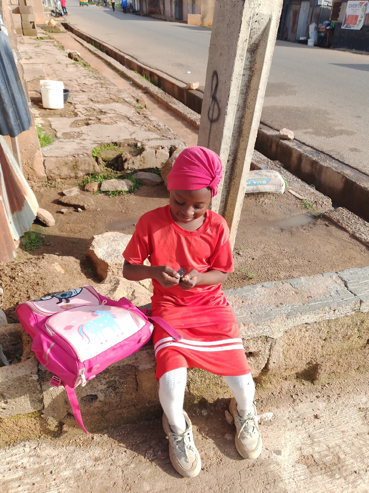 In Jos, Nigeria, a young girl wearing a pink headscarf and a red t-shirt sits on a weathered concrete ledge. She has white socks and beige sneakers, and a bright pink backpack with a unicorn design rests beside her. She is looking down, engaged with a small object in her hands. The environment is dusty with scattered rocks and a partially visible street with some traffic in the background, suggesting an outdoor urban setting. A concrete pillar stands nearby, bearing graffiti.