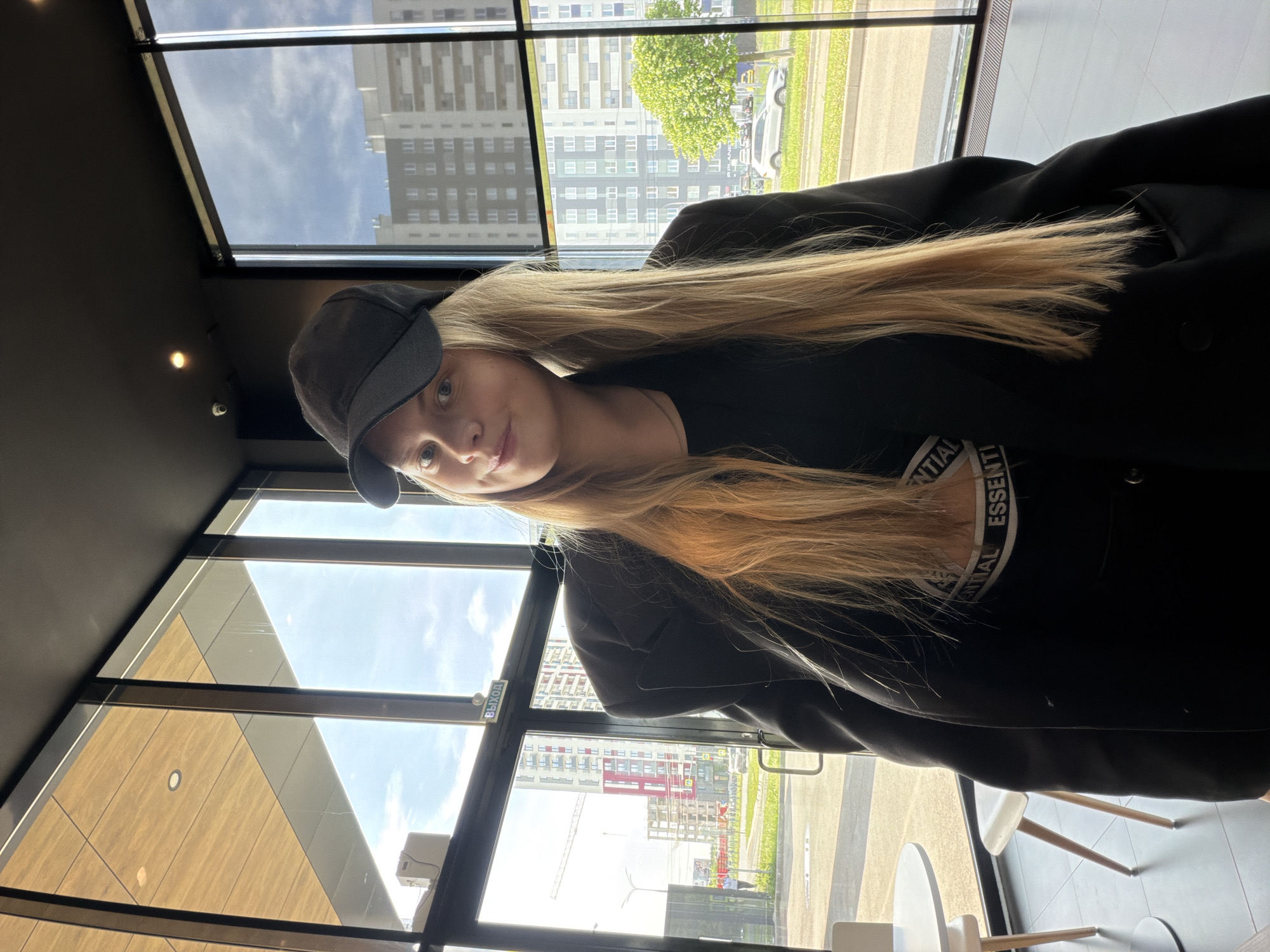 The image shows a woman with long blonde hair and wearing a black cap, black t-shirt with a white graphic, and a black jacket. She is inside a building, likely a cafe or restaurant, with large windows looking out onto a cityscape. Visible through the windows are tall apartment buildings, trees, and a street with pedestrian walkways.
Based on visual cues, the scene is located in Izhevsk, Russia. In particular, the style of apartment buildings and the road markings are consistent with that region. The bright daylight indicates it is likely daytime. The woman is looking directly at the camera with a slight smile. The overall setting suggests a casual, indoor scene in a modern building in an urban area.