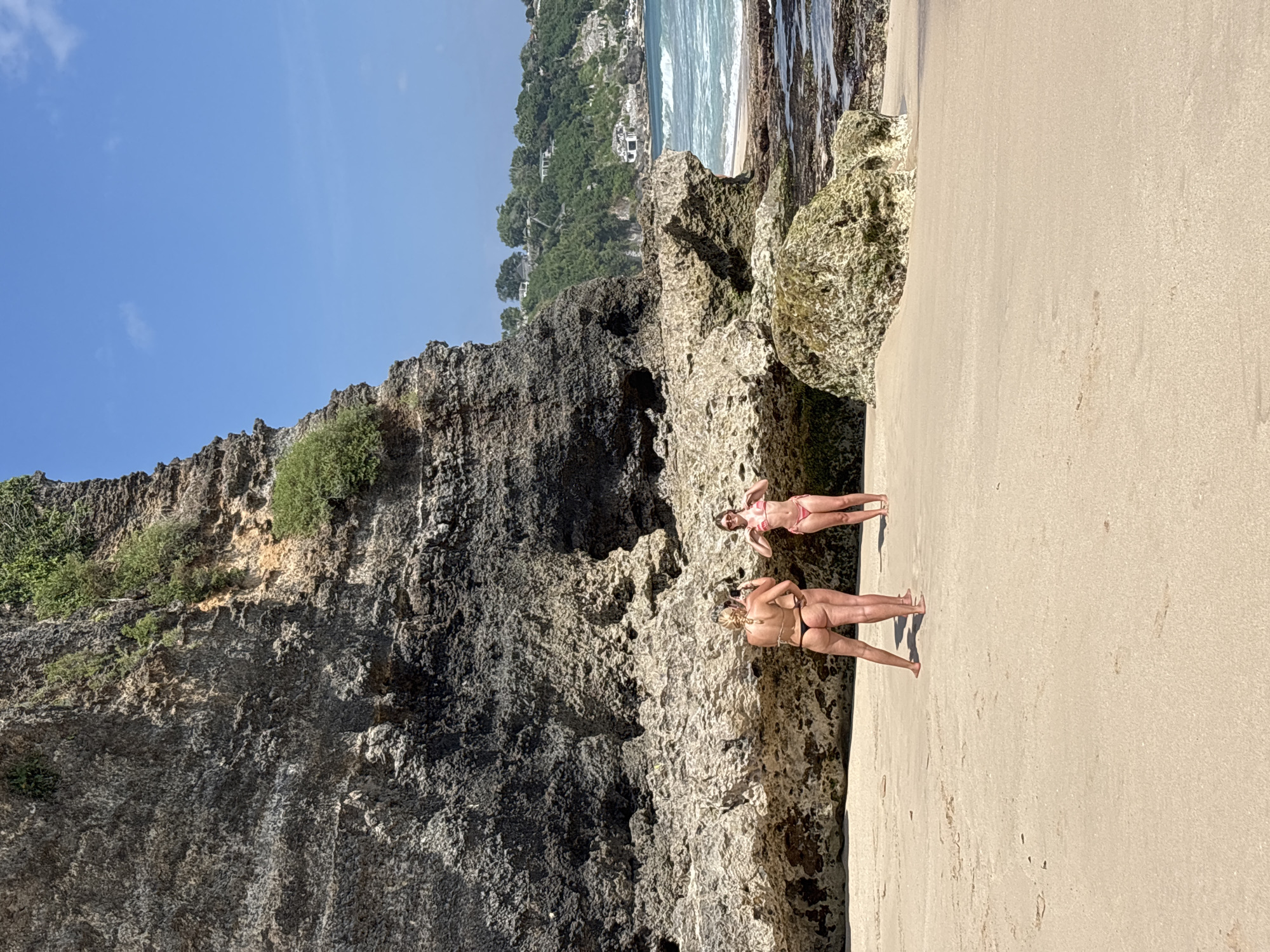 In Kuta Selatan, Indonesia, two women stand on a sandy beach. One woman, facing away from the camera, wears a black bikini and has blonde hair styled in a braid. The other woman, also facing away, wears a pink bikini and sunglasses. They are positioned near a large, rocky cliff face that dominates the left side of the frame. The cliff is dark grey and covered in patches of green vegetation. In the background, the turquoise ocean meets a lighter blue sky. Buildings and lush green hills are visible in the distance, hinting at a developed coastal area. The sand is light brown and appears relatively smooth, with some footprints and small debris scattered across it. The water exhibits gentle waves, and rocky outcroppings are present near the shore.