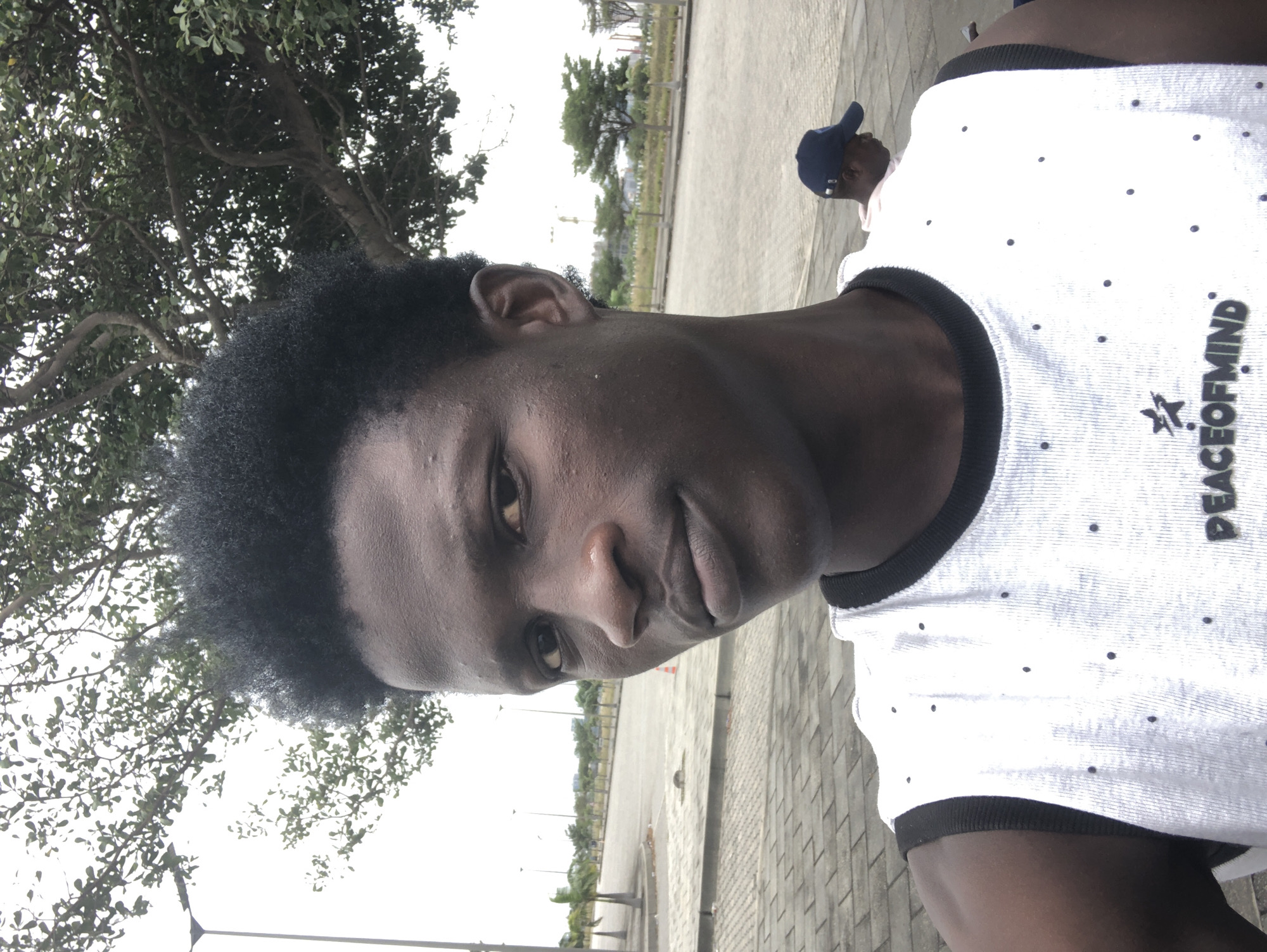 A close-up, selfie-style shot features a young Black male facing the camera. He has dark skin, short, dark, curly hair, and is wearing a white, sleeveless shirt with black trim. The shirt has a pattern of small black dots and text that reads "PEACEOFMIN" followed by a star graphic. The background shows a paved area with trees and streetlights, suggesting an outdoor urban environment. In the mid-ground, another person wearing a dark blue baseball cap is visible. The lighting is bright, indicating daylight. The overall scene appears to be an open, possibly public space in Ikoyi, Nigeria.