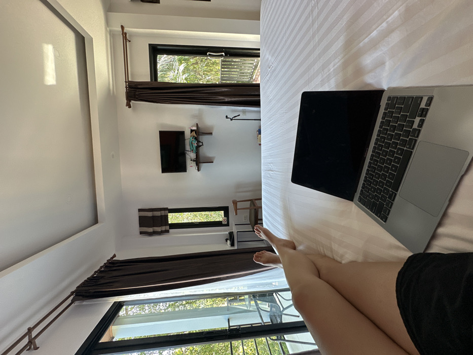 A forensic description of the scene in Phra Ae, Thailand, reveals an indoor room, likely a hotel or resort accommodation.
**People:**
One individual is present, with only their lower body (legs and feet) visible, lying prone on a large bed. They are wearing dark shorts.
**Subjects/Objects:**
A silver Apple MacBook laptop with a dark screen is positioned on the white, horizontally striped bedding in the foreground. The bed occupies the lower half of the frame. The room features white walls and a white ceiling with a rectangular recessed panel design.
On the left, a large glass sliding door with a black frame is partially open, revealing a balcony with a black metal chair and table. A brown curtain hangs to the side of this door, with a sheer white curtain also present. To the right of this, a tall, narrow window with a brown and white striped roman blind is visible. Below it, a dark side table is present.
A flat-screen television is mounted on the wall above a dark wooden floating shelf, which holds several unidentified objects. A light-colored wooden chair is situated in the corner below the TV. A black monopod or tripod stands vertically on the floor to the right of the TV shelf. Another large glass sliding door with a black frame and a brown curtain is on the right, showing an exterior with lush green foliage and a wooden slatted railing.
**Actions/Interactions:**
The individual is resting on the bed while the laptop is positioned open, suggesting potential use or preparation for use. Natural light illuminates the room through the open doors and window, revealing a verdant outdoor environment.
**Environment:**
The interior appears clean and modern. The exterior view indicates a tropical or subtropical climate, consistent with Thailand.