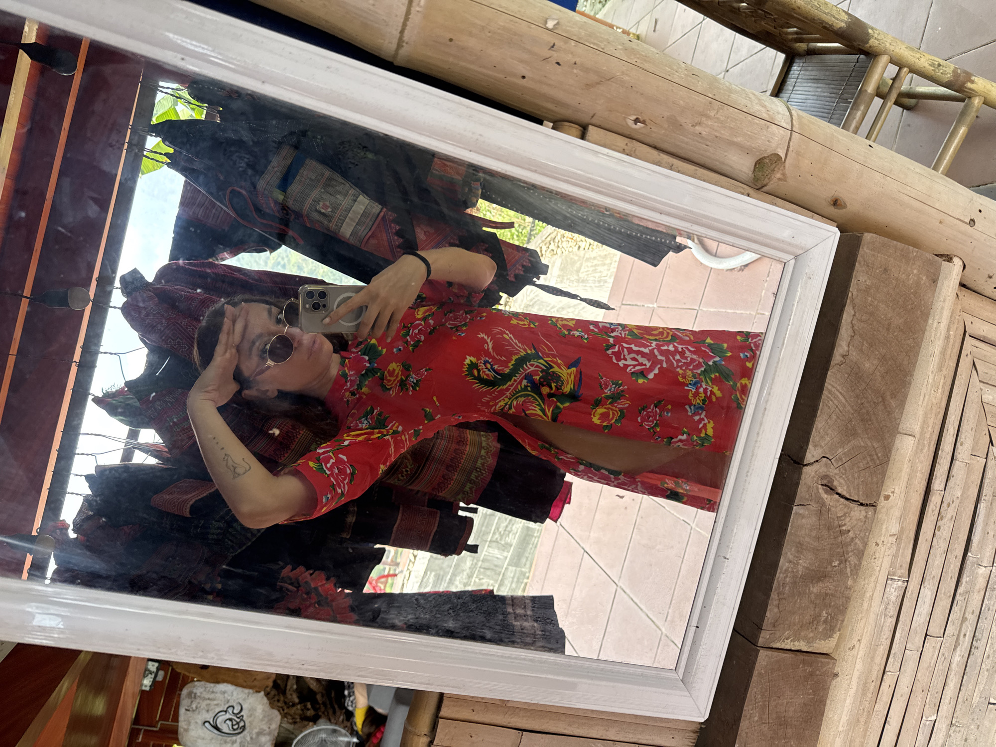The image shows a woman taking a mirror selfie in what appears to be a shop or guesthouse in Thông Nguyên Commune, Vietnam. The woman is reflected in a large, white-framed rectangular mirror, seen from the waist up. She is wearing a vibrant red dress adorned with a floral pattern and a golden dragon motif. She has dark, round sunglasses on, and her left hand is casually placed near her head. In her right hand, she holds a light-colored smartphone with a prominent camera array (likely an iPhone), pointing it towards the mirror to capture the selfie. A small, stylized tattoo, possibly depicting an animal, is visible on her left forearm.
Behind the woman's reflection, numerous colorful, patterned textiles, resembling traditional Hmong clothing or fabric, are hanging, suggesting they are merchandise or part of the decor. Beyond these textiles, a bright outdoor scene with clear skies and some foliage is visible, indicating it is daytime.
Outside the mirror's frame, the surrounding environment features natural wood and bamboo elements. A large, light-colored bamboo log forms part of a structure, possibly a bed or bench, positioned on a tiled floor. Additional bamboo poles create a textured surface, either as flooring or a wall element. The rustic bamboo construction and the traditional textiles visible in the reflection strongly support the location context of rural Vietnam. The overall atmosphere appears bright and casual. No discernible text is visible apart from the phone's design elements.