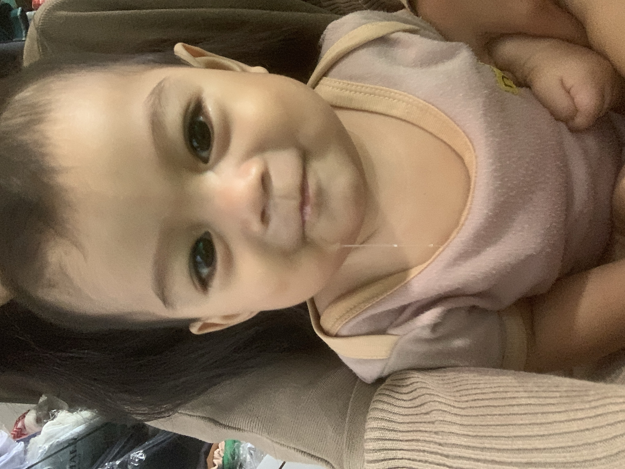 A close-up, eye-level, head-and-shoulders portrait shows a baby with dark hair, dark eyes, and a light complexion. The baby is wearing a light pink onesie with tan trim. A string of saliva hangs from the baby's chin. The baby is being held by an unseen adult, as indicated by the presence of adult clothing and hair in the background. The image appears to have been taken indoors, possibly in a home environment. There are no distinguishing geographical markers to place the image specifically in Sungai Meriam, Indonesia, or any other city.