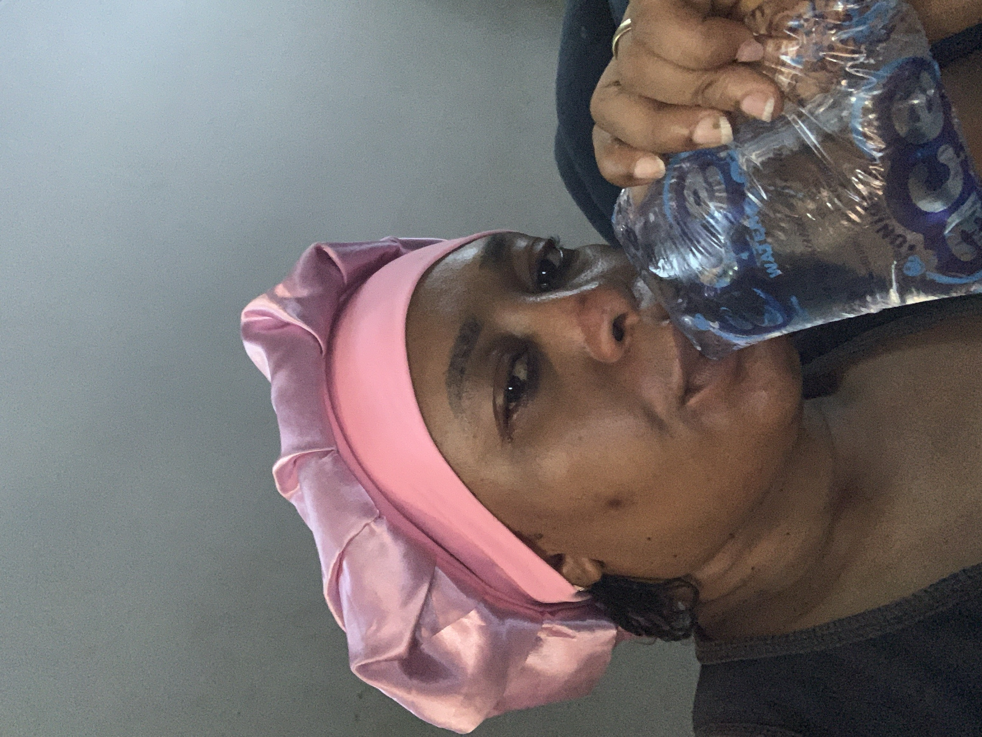 A person, presumably a woman, is drinking water from a plastic bottle. She is wearing a pink satin head covering. The location is unknown, so I cannot confirm if it is in Sabon Tasha, Nigeria.