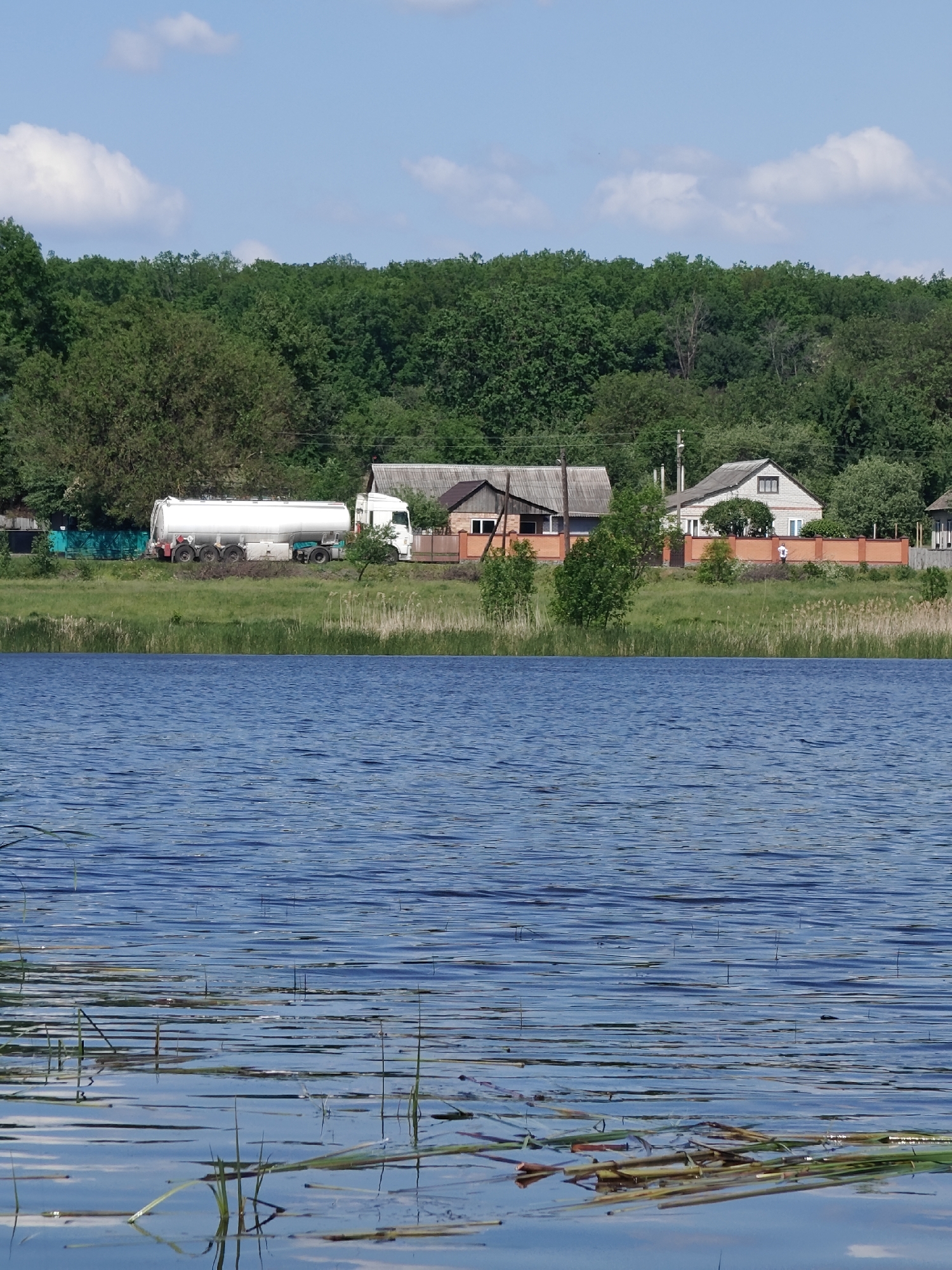 The image shows a lake with vegetation in the foreground. Across the lake, there is a grassy bank with buildings. A white tanker truck is parked in front of the buildings. Behind the buildings, there is a line of trees. The sky is blue with some white clouds. The location is Bobrivnyk, Ukraine.