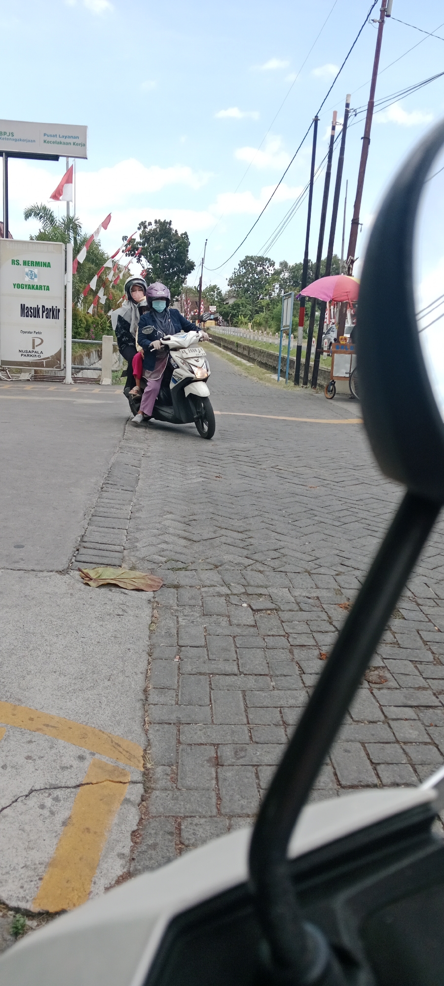 This is a vertical, eye-level, medium shot of two individuals on a white scooter in Maguwoharjo, Indonesia. The scooter is moving forward on a brick road. The person in front is driving, wearing a dark blue jacket, light purple pants, a light purple helmet, and a light blue face mask. They are wearing white gloves. The person behind is wearing a black jacket, pink pants, and a white helmet with a light blue face mask. The scooter has a license plate that reads "AE 2868 AV".
To the left of the road is a sidewalk with yellow markings. A sign reads "RS. HERMINA YOGYAKARTA". Further left, another sign reads "BPJS Ketenagakerjaan" and "Pusat Layanan Kecelakaan Kerja". Two flags, one red and white, and another white with red stripes, are visible.
To the right of the road, there are utility poles with wires. A pink umbrella is set up, and a cart is nearby. Trees and foliage are visible in the background. The sky is blue with some white clouds. The image appears to be taken from another vehicle, as the side mirror of a vehicle is visible in the foreground.