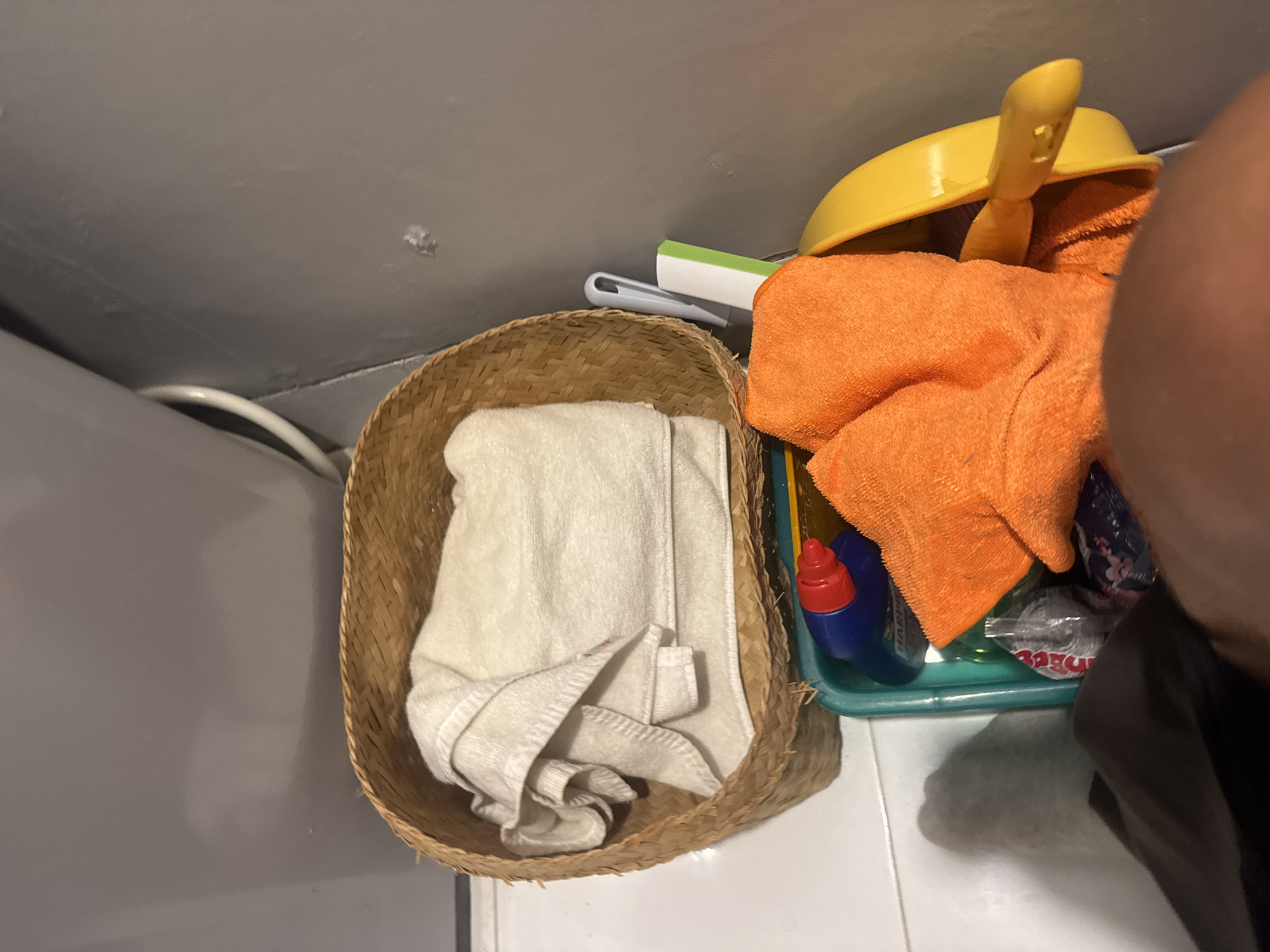 A woven basket sits on a tiled floor, containing a folded white towel. To the right of the basket is a green plastic tray filled with cleaning supplies. A bright orange towel is draped over some of the items in the tray. A yellow dustpan with a handle is resting on top of the orange towel. Partially visible in the tray are a blue bottle with a red cap, a green bottle, and a clear plastic package with red lettering. The scene appears to be in a bathroom or utility area, indicated by the tiled floor and the presence of cleaning products. The lighting suggests it is daytime, and there are no explicit indications of weather. The overall impression is a tidy, organized storage space for cleaning items. The lettering on the clear plastic package in the tray is partially visible and appears to be in English, possibly a brand name or product description.