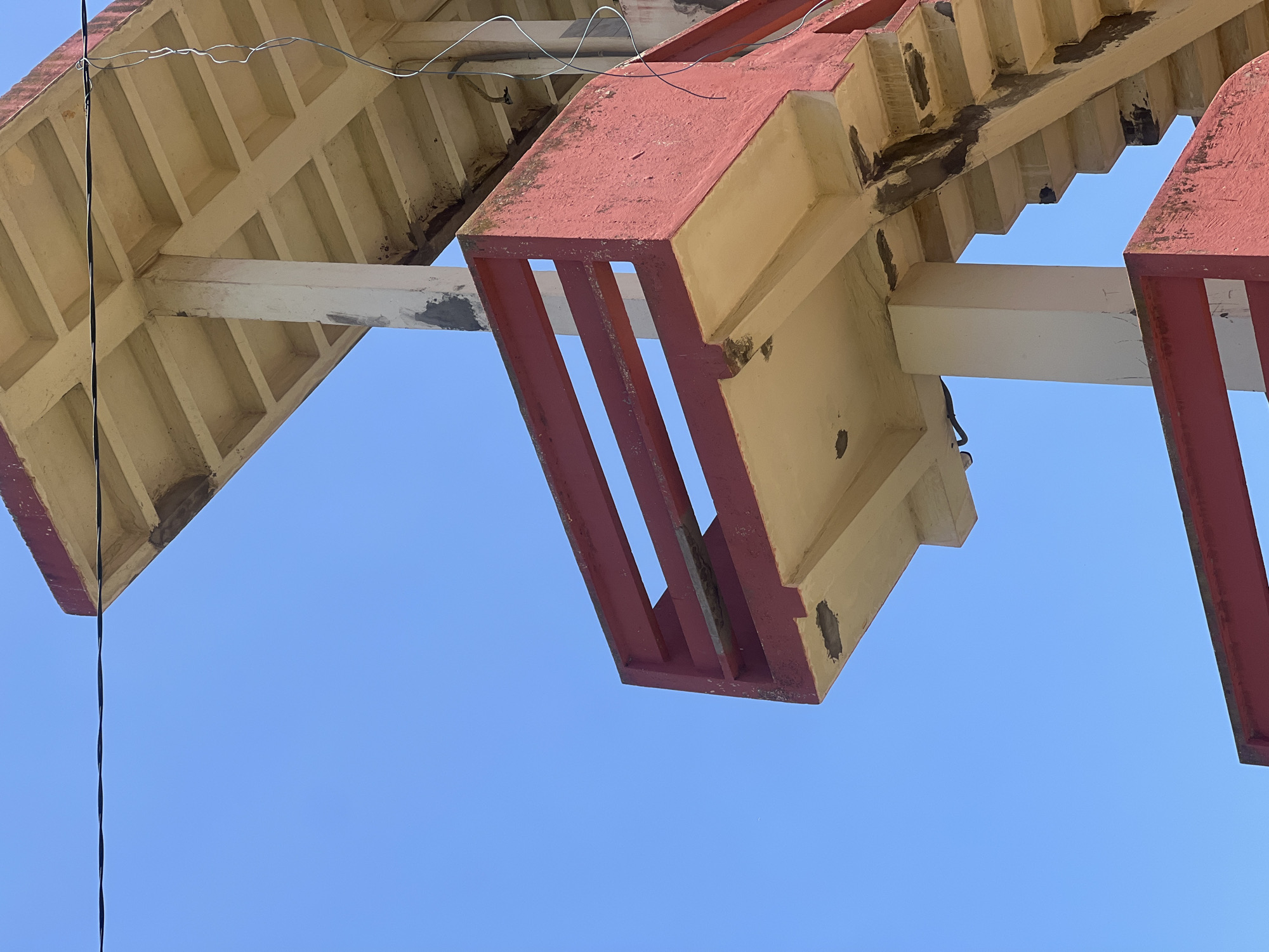 The image displays an upward-angled view of a multi-level, weathered concrete structure set against a clear, light blue sky. The structure features prominent horizontal railings and some structural elements painted in a faded, muted red, while the undersides of platforms and the visible staircase are a light beige or cream color.
The upper section of the structure shows a flat platform with a distinctly coffered (grid-patterned) underside in beige, supported by thick, rectangular white columns, which exhibit peeling paint and discoloration. A thick, black electrical cable runs horizontally across the upper third of the frame, passing near the top platform. From the right side of the upper platform, a thinner, wavy grey wire dangles vertically.
Below the top platform, a middle level features a section with a three-bar horizontal railing, also in muted red, extending towards the left. This platform's underside is beige and shows significant signs of wear, including dark grey patches indicative of repairs or sealant applications. To the right, a concrete staircase ascends, revealing similar dark grey repairs along its risers and underside. The overall condition of the structure suggests age and exposure to elements, with widespread fading, chipping, and cracks on painted and unpainted surfaces.
No people, vehicles, vegetation, or ground features are visible. The clear blue sky provides the sole background. While the structure's design and materials are consistent with coastal or public infrastructure found in cities, the image itself does not contain specific identifying markers for a precise location within Casablanca, Morocco.