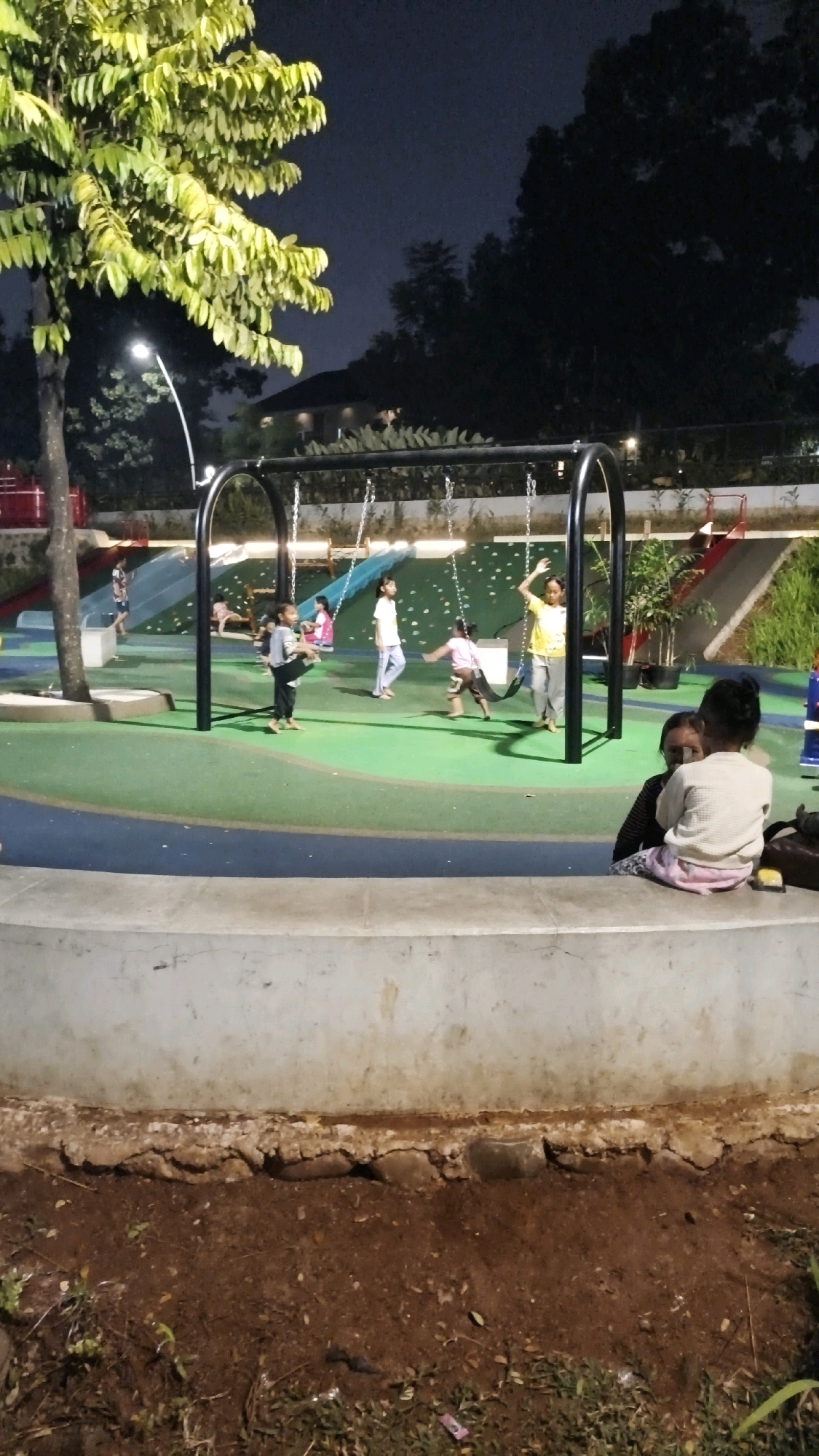 The image depicts a nighttime scene at a playground in South Jakarta, Indonesia. Several children are engaged in various activities on the playground. Two children are on swings, with one appearing to be mid-swing and the other standing beside the swing set. Another child is walking across the green play surface, and two more children are visible in the background near a slide. In the foreground, two adults and a child are seated on a concrete ledge, with one adult and the child looking towards the playground. The playground features a green and blue rubberized surface, a swing set, slides, and surrounding greenery. The lighting suggests it is after sunset, with artificial lights illuminating the playground.