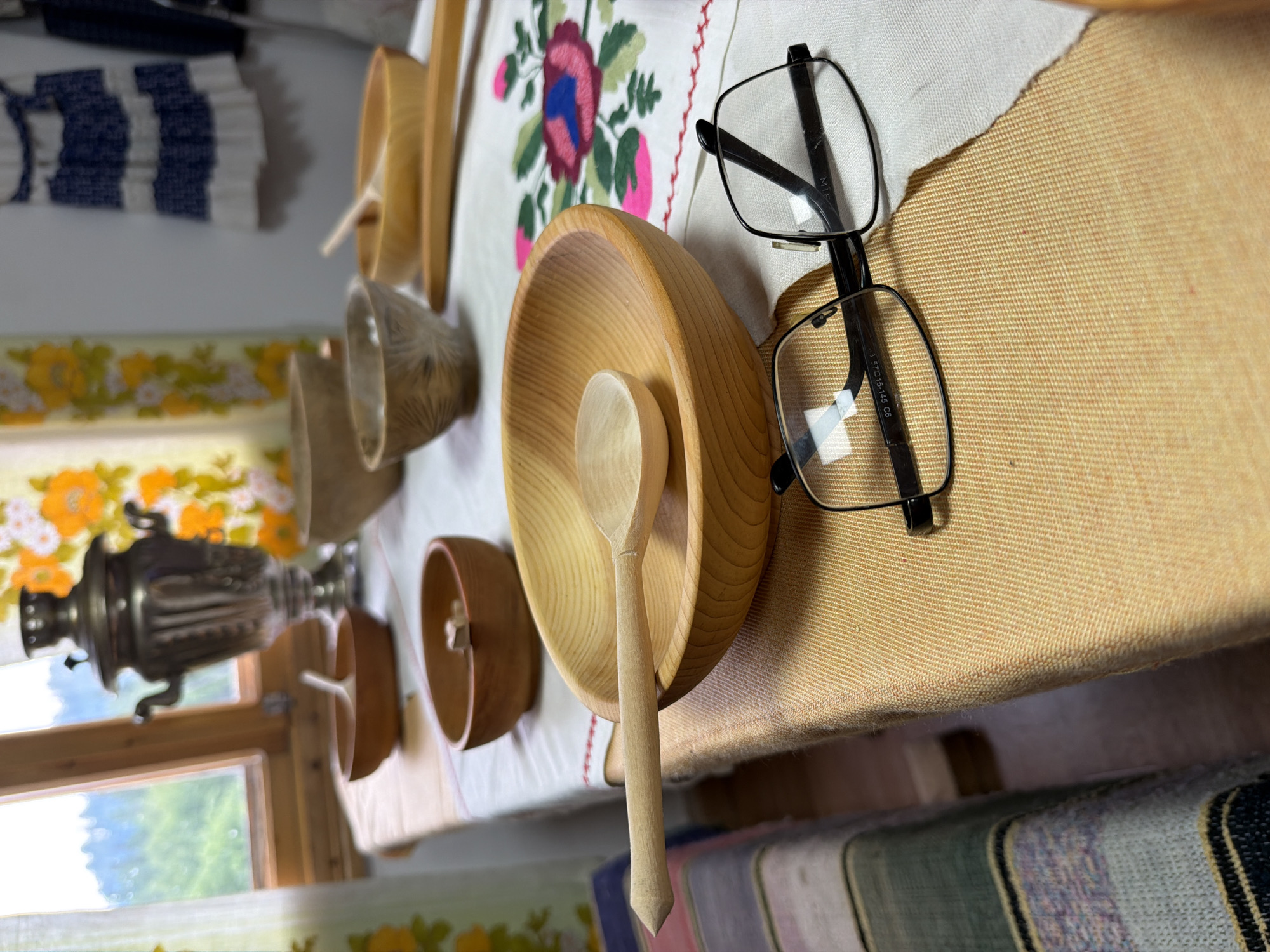 An indoor scene depicts a wooden table set with several handcrafted wooden bowls and spoons, a traditional Russian samovar, and a pair of eyeglasses. The table is covered with a tablecloth featuring embroidered flowers. The background reveals a window with a view of a green, mountainous landscape. The overall impression is one of a rustic, home setting, likely in a rural area, possibly a guesthouse or a domestic space in Ust'-Kabyrza, Russia. The lighting is natural, coming from the window.