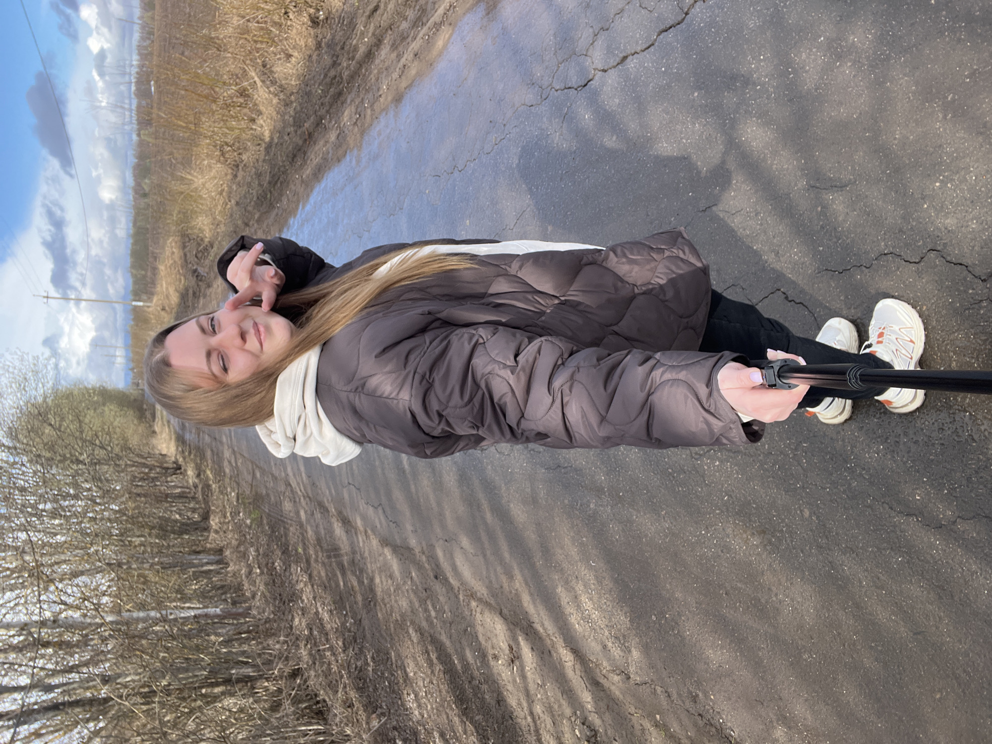 A young woman with long, light brown hair is posing for a selfie on a cracked asphalt road in Opalevo, Russia. She is wearing a dark brown puffer jacket over a light-colored hoodie, black pants, and white sneakers with orange accents. She is holding a black selfie stick. The road appears slightly wet and runs alongside a field with bare trees and vegetation. The sky is visible in the distance, with patches of blue and white clouds.