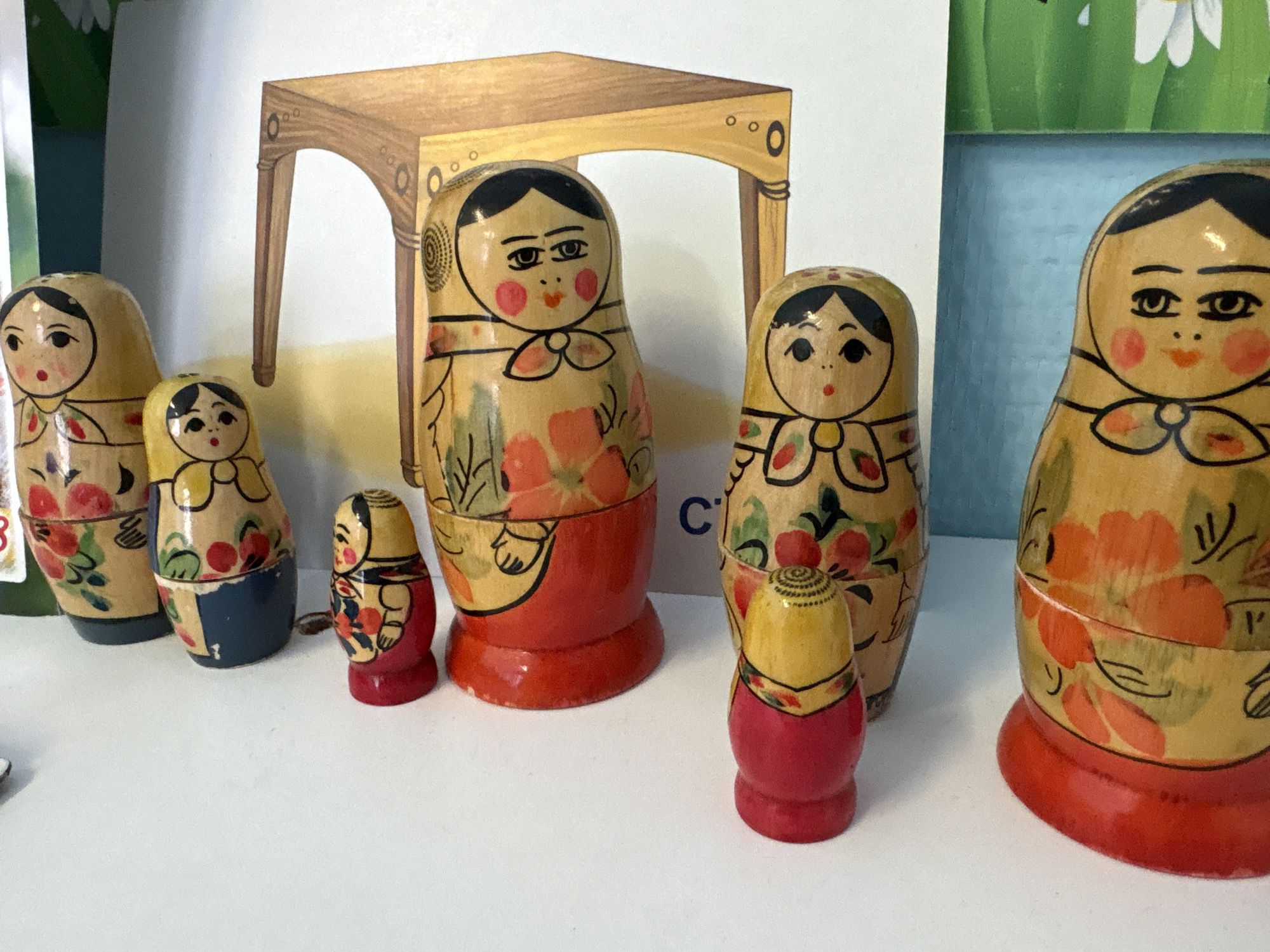 The image presents seven matryoshka dolls of varying sizes, arranged in a linear configuration on a flat, white surface, located in Chaikovsky, Russia. Each doll features a light wood-toned body with painted black hair, facial details (eyes, eyebrows, nose, mouth), and red cheeks. The dolls are decorated with floral patterns primarily in red, orange, and green hues. Five dolls have red-painted bases, while two smaller dolls on the left have dark blue bases. The smallest two dolls, positioned near the center and right, feature yellow-painted tops and red bases. The doll furthest right is partially cropped.
The background consists of a multi-sectioned illustration. On the left, green foliage and a white flower are visible. Centrally, a light brown illustration of a wooden table or bench with two visible legs and decorative circular motifs is present. A blue capital letter "C" is visible on the background to the right of the central large doll. A segment of light blue background is also visible towards the far right. No people or actions are depicted in the scene.