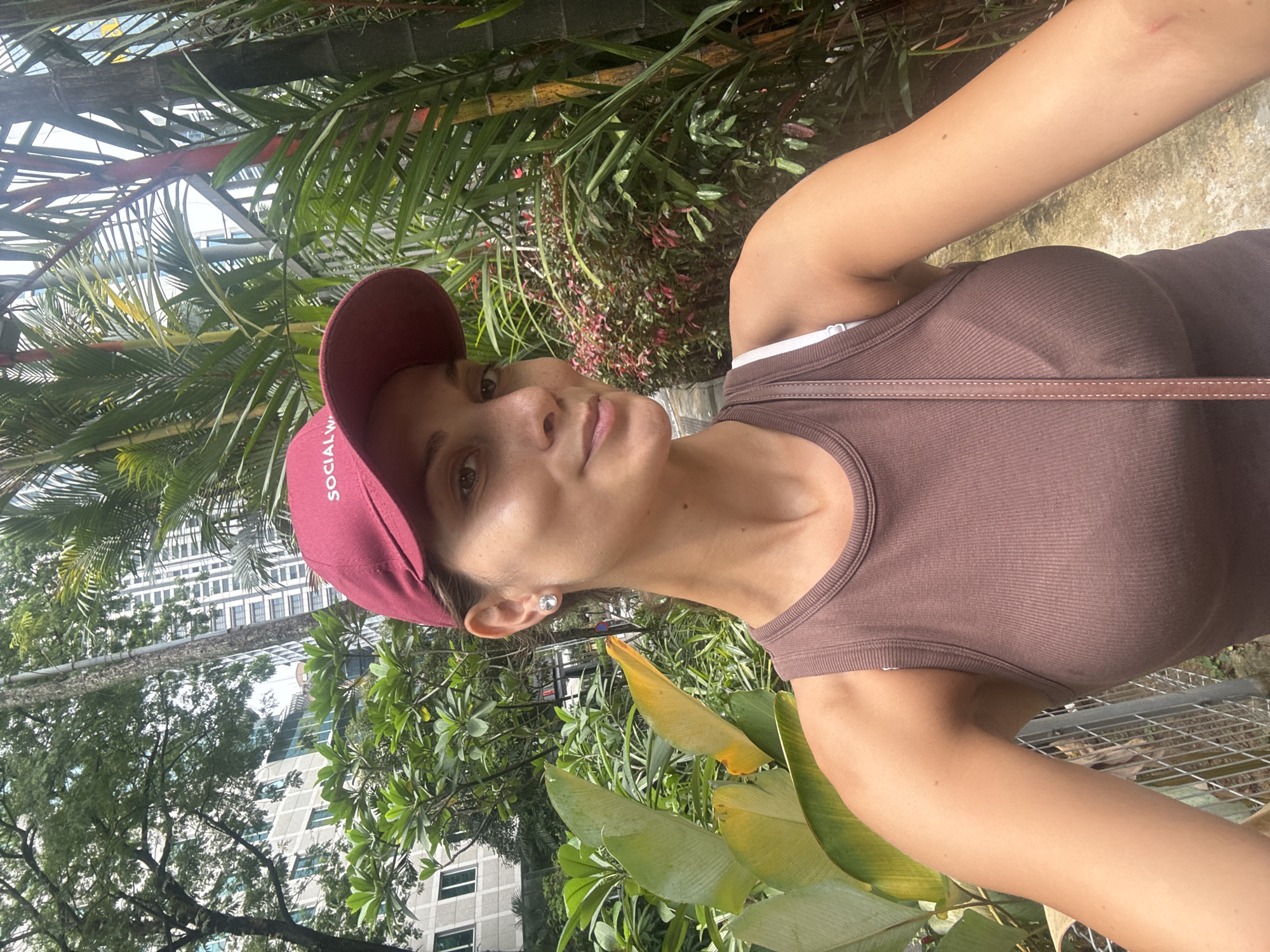 A woman stands in the KLCC Park in Kuala Lumpur, Malaysia. She is wearing a maroon baseball cap, a brown tank top, and a crossbody bag. She is surrounded by tropical plants. Skyscrapers are visible in the background. The photo appears to be a selfie.