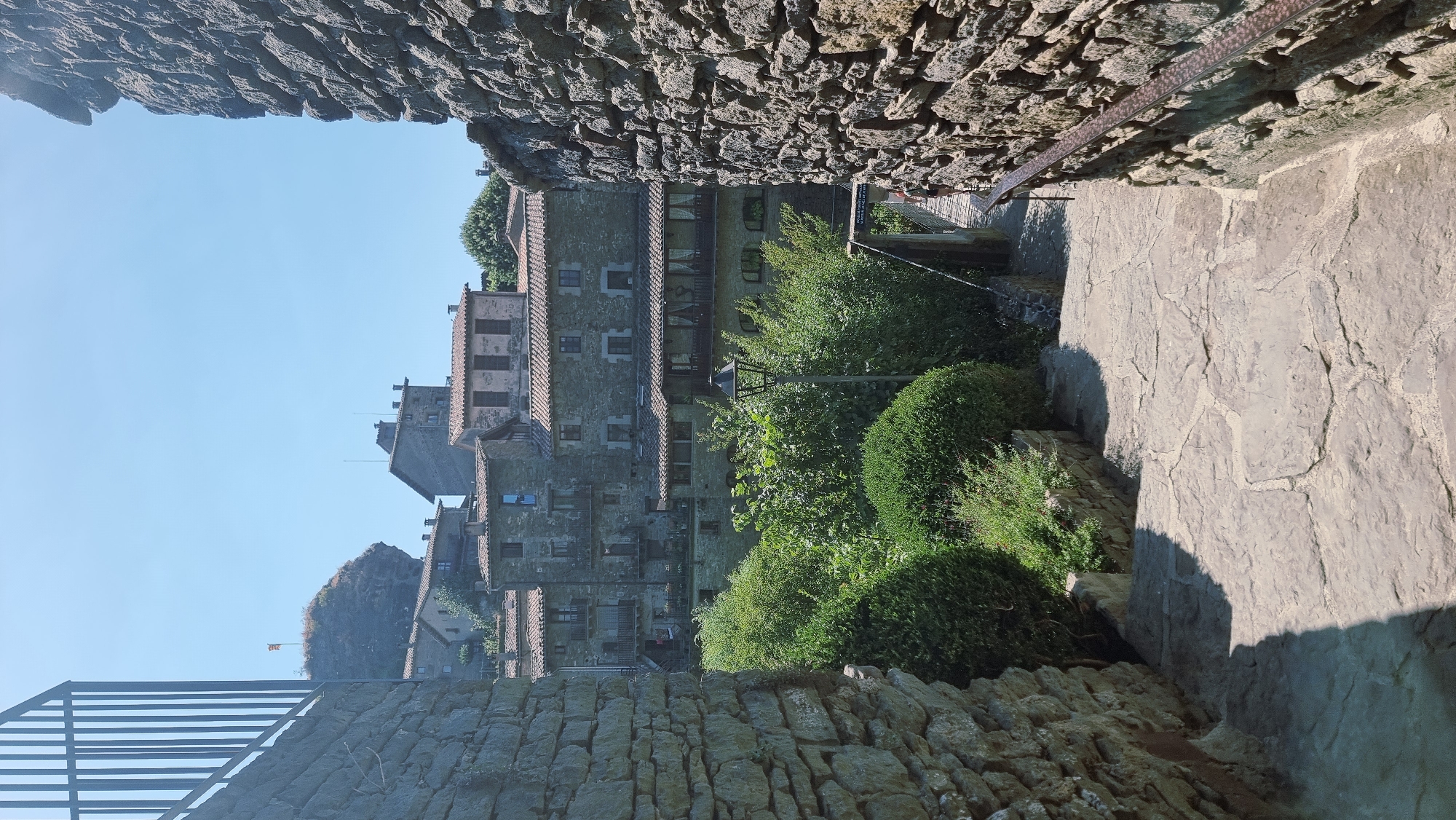 The image captures a sunlit, stone-paved pathway descending between two weathered stone walls in the medieval village of Rupit i Pruit, Spain. The path, made of irregular flagstones, is bordered by neatly trimmed green bushes on the left and a rustic metal handrail along the stone wall on the right.
In the background, a cluster of traditional stone houses with terracotta roofs are visible, stacked on a hillside. These buildings are characterized by their aged stone facades, wooden balconies, and small windows. A flag is visible atop a prominent rocky outcrop in the distance. A vintage-style streetlamp stands amidst the greenery. The scene is bathed in bright daylight, casting distinct shadows. The sky is clear and blue.