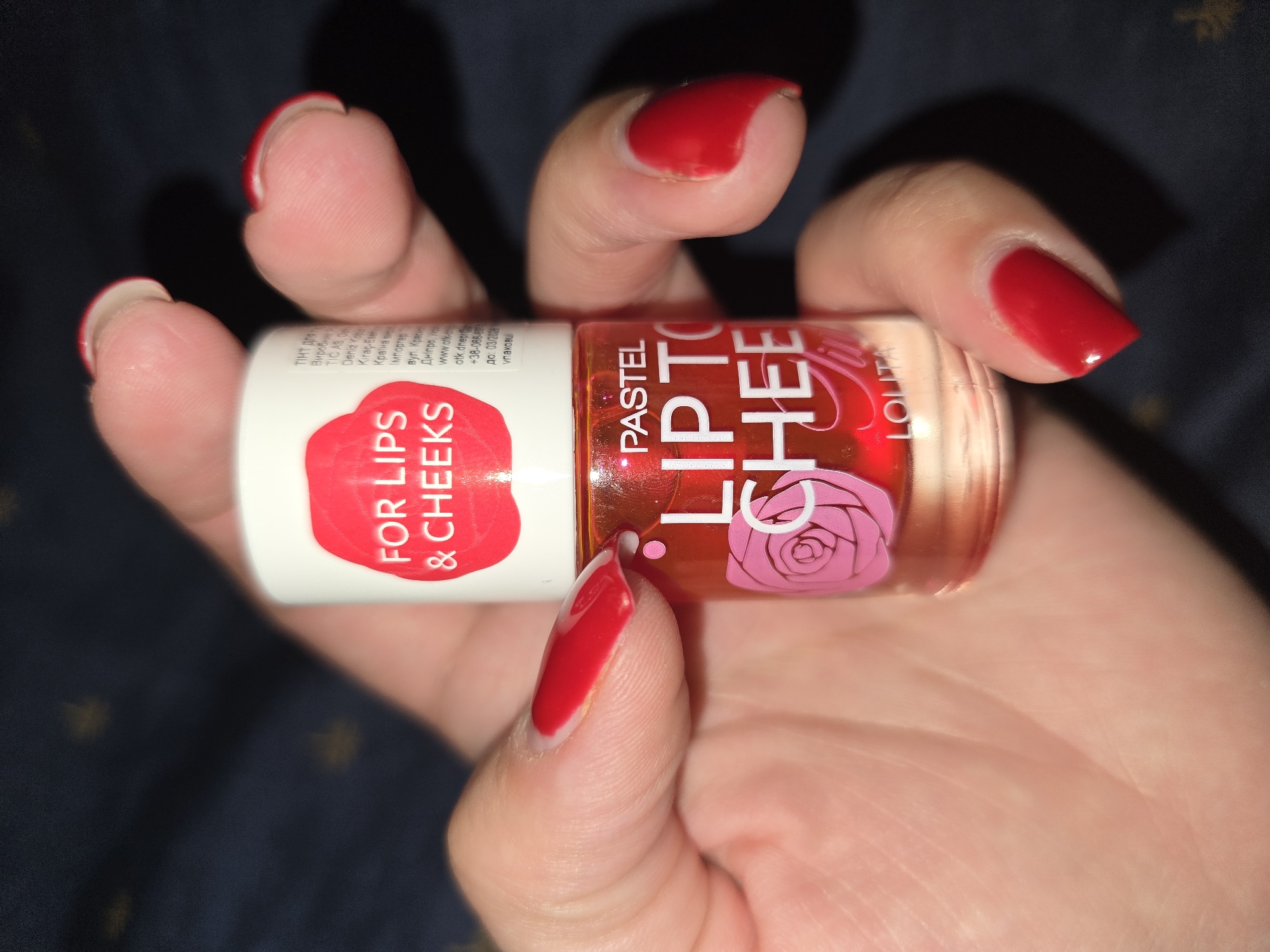 A person's hand with red nail polish holds a small bottle of "PASTEL LIP TO CHEE" cosmetic product. The bottle is transparent and filled with a red liquid. The label on the bottle has text in English and possibly another language, with a pink rose graphic. The background is dark and appears to be a fabric with a subtle pattern. The image is a close-up shot of the hand and the product. There is no information in the image that suggests the location is Petropavlivska Borshchahivka, Ukraine.