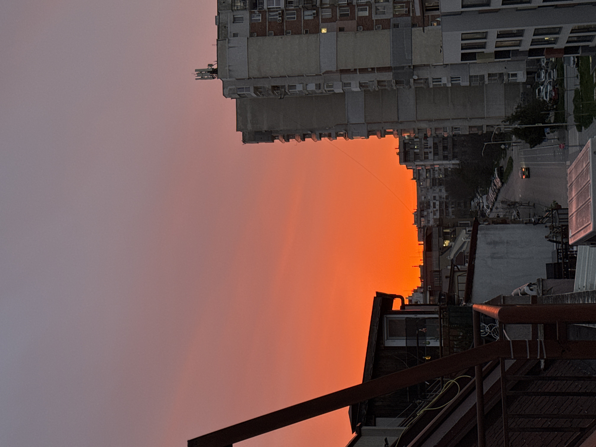 The image captures a sunset in Varna, Bulgaria, from a high vantage point. The sky is ablaze with orange hues near the horizon, transitioning to a softer pink and gray towards the upper left. The foreground features architectural elements, including metal stairs and rooftops. In the mid-ground, the scene shows a densely populated urban area with tall apartment buildings. A street lined with parked cars is visible. The buildings are constructed of pre-cast concrete.