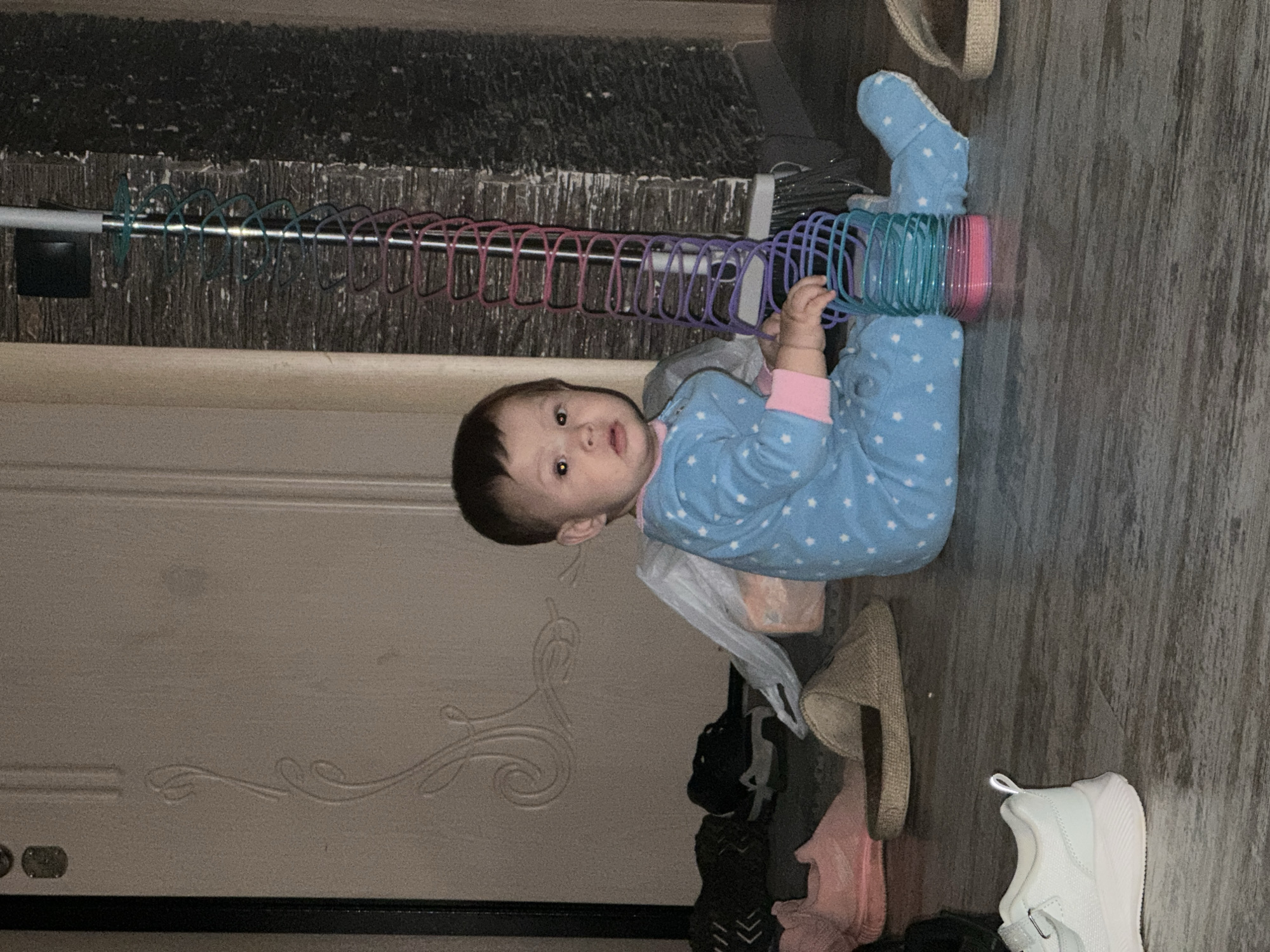 An infant wearing a blue pajama set with white stars sits on a wooden floor next to a broom with a colorful, coiled toy attached to its handle. The child is positioned on the right side of the image, facing forward with a neutral expression. To the left of the child, several pairs of footwear are scattered on the floor, including pink slip-on shoes, a pair of black sneakers, and a brown slipper. A white shoe is visible in the foreground on the left. Behind the child is a portion of a white door with decorative carvings. The environment appears to be indoors, possibly a hallway or entryway. The lighting is dim, creating shadows and a grainy texture in the image.