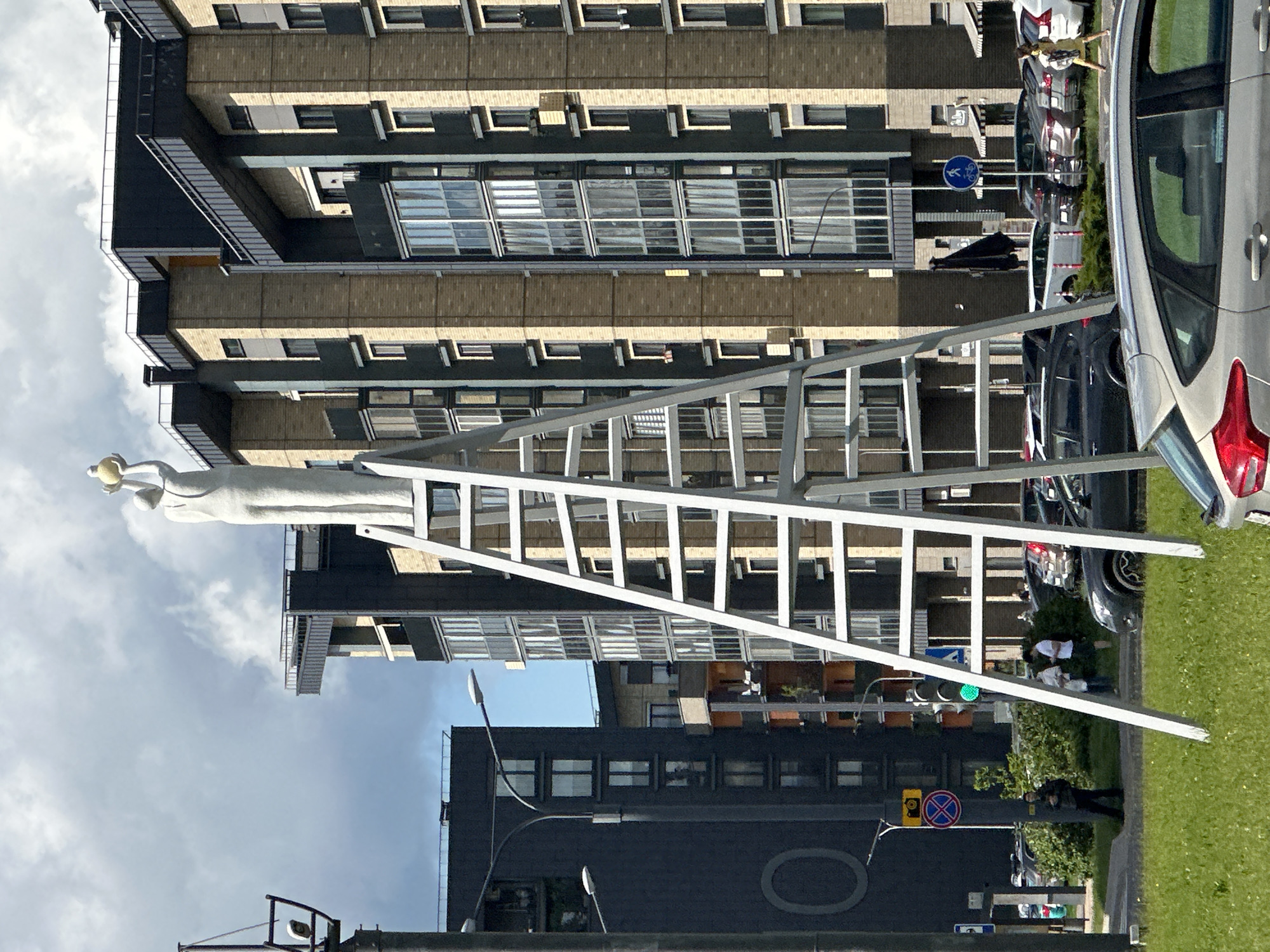 The image, taken in Бараўлянскі сельскі Савет, Belarus, shows a street scene dominated by a tall apartment building. In the foreground, a ladder-like structure is angled across the view, partially obscuring the buildings behind it. A statue of a figure in white is visible on the left side of the ladder structure.
The apartment building features horizontal rows of windows, and architectural elements give it a layered appearance. A black building with a large oval shape on its side is visible below.
People are scattered along the sidewalk near parked cars. Traffic lights, street signs, and greenery add to the street scene. The sky is cloudy.
