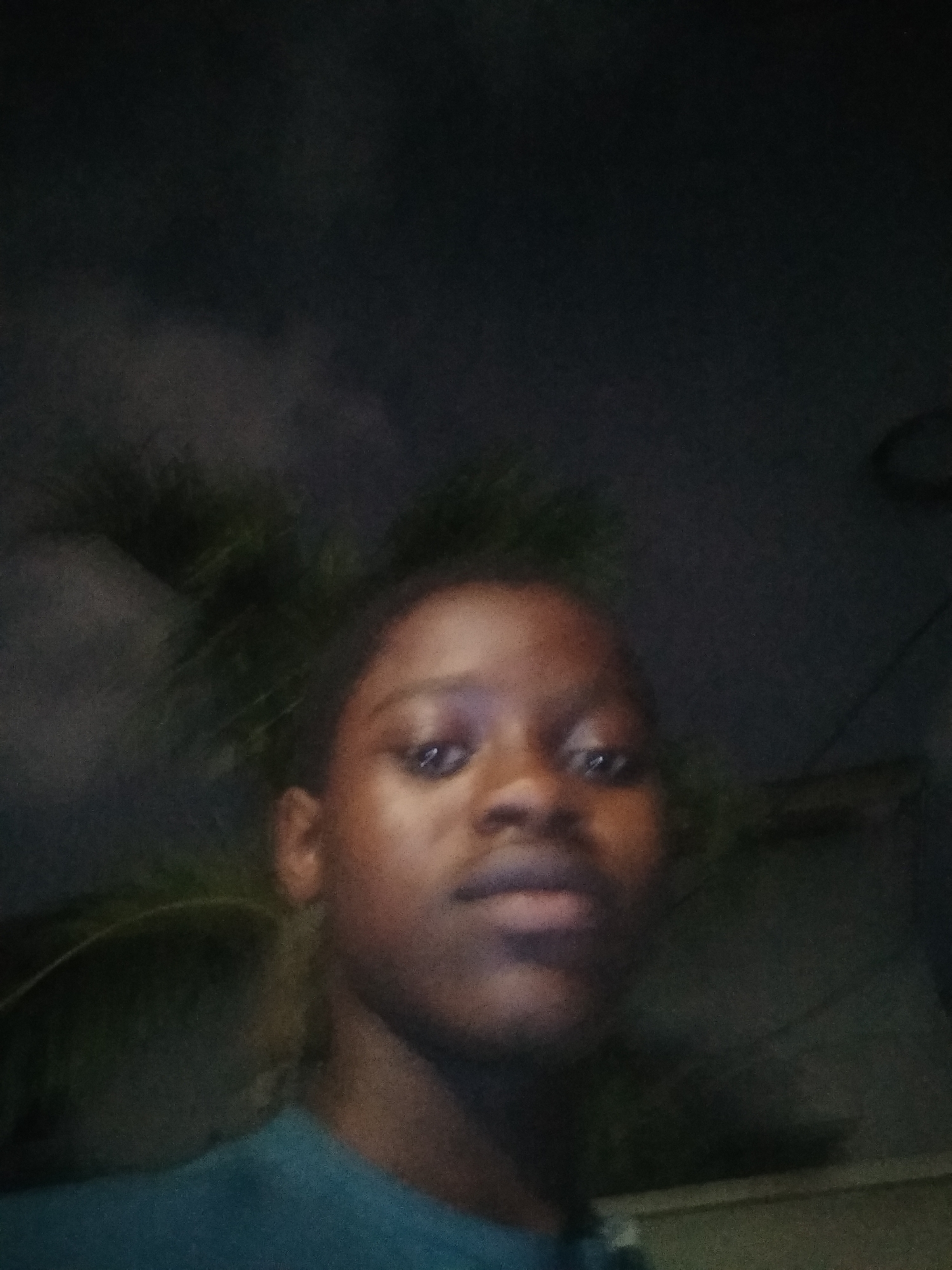 A close-up, low-light photograph features a dark-skinned person centered in the frame. The individual appears to be outdoors at night in Shangisha, Nigeria, indicated by the presence of palm trees and the low ambient light. The background is dark, suggesting a nighttime or dimly lit environment. The person is wearing a dark-colored shirt. The focus is on the face, but it is somewhat blurry due to the lighting conditions. There is no visible interaction or action taking place.