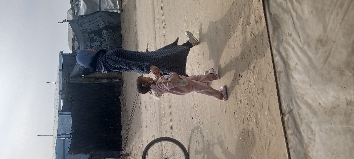 A woman in a dark blue patterned dress and light blue headscarf holds a small child in a pink outfit. They stand on a sandy ground near makeshift shelters, with a bicycle wheel visible nearby. The scene suggests a refugee camp or temporary housing situation, under a muted sky.