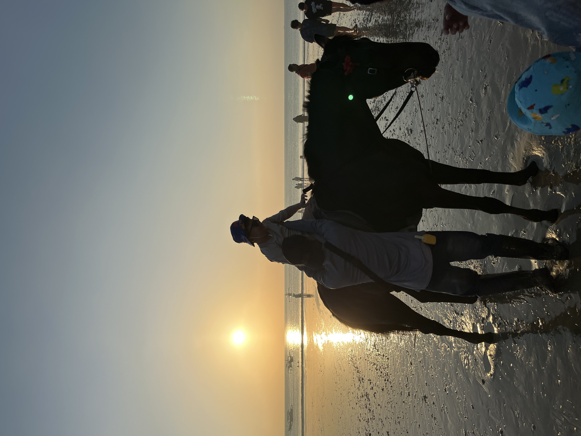 This image captures a serene sunset scene on the beach in Kuta Selatan, Indonesia. In the foreground, a dark-colored horse stands on the wet sand, with a person, likely the rider's parent or handler, holding onto the bridle. A child, wearing a hat and sunglasses, sits atop the horse. Further back, several other people are seen strolling along the shoreline and wading in the shallow water. Traditional Balinese boats are visible on the calm ocean surface, and the setting sun casts a warm, golden glow across the sky and reflects on the water, creating a tranquil atmosphere. The sand is wet and speckled with small shells and debris, indicating the receding tide.