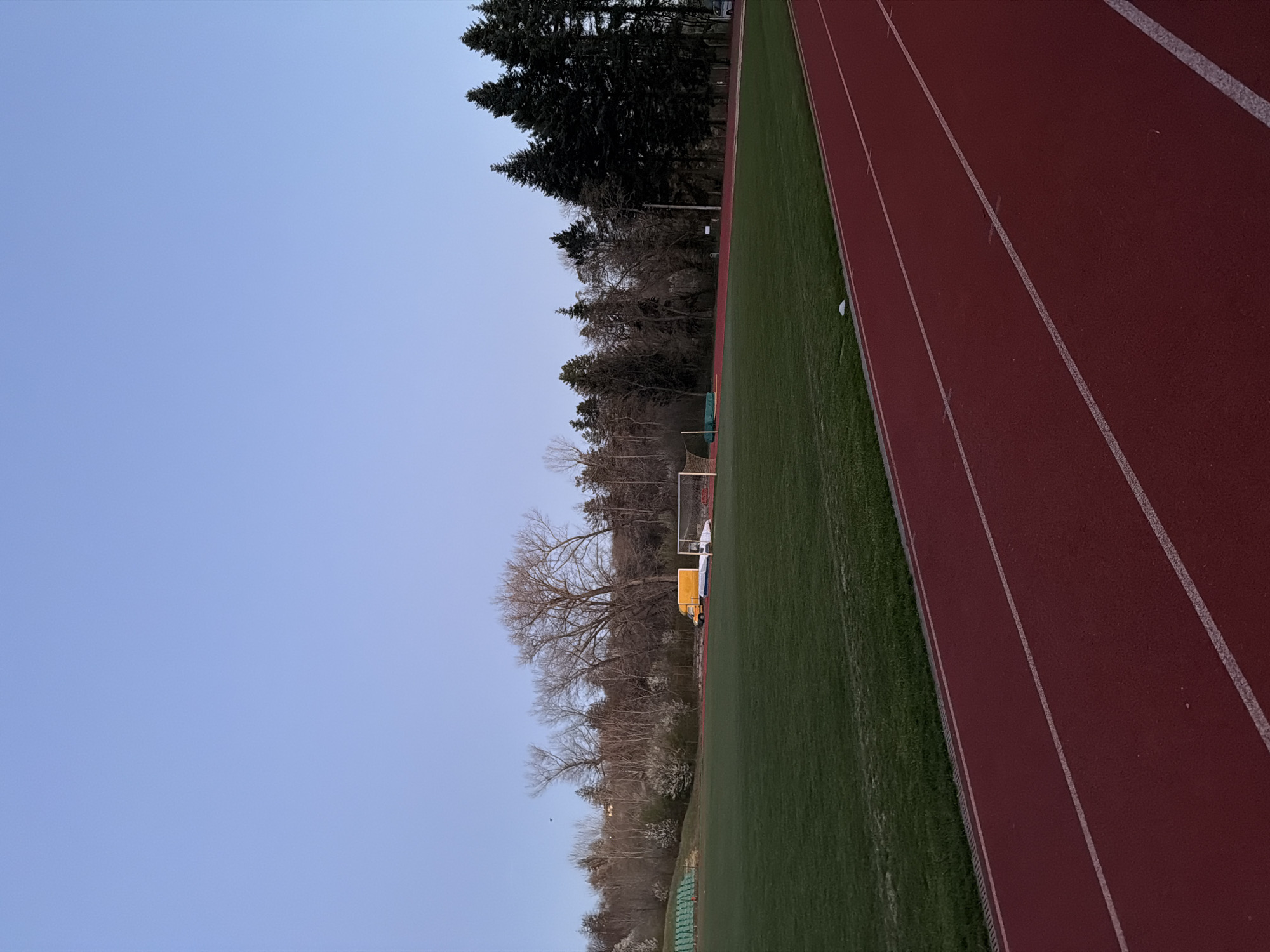 The image depicts a track and field area adjacent to a soccer field, with a running track on the right, green turf in the middle, and a backdrop of trees on the left. In the background is a soccer goal. There is a yellow vehicle near the soccer goal. The sky is a pale blue. Based on the type of track, location, and proximity to tree, this image is possibly from the Panagyurishte Stadium in Panagyurishte, Bulgaria.