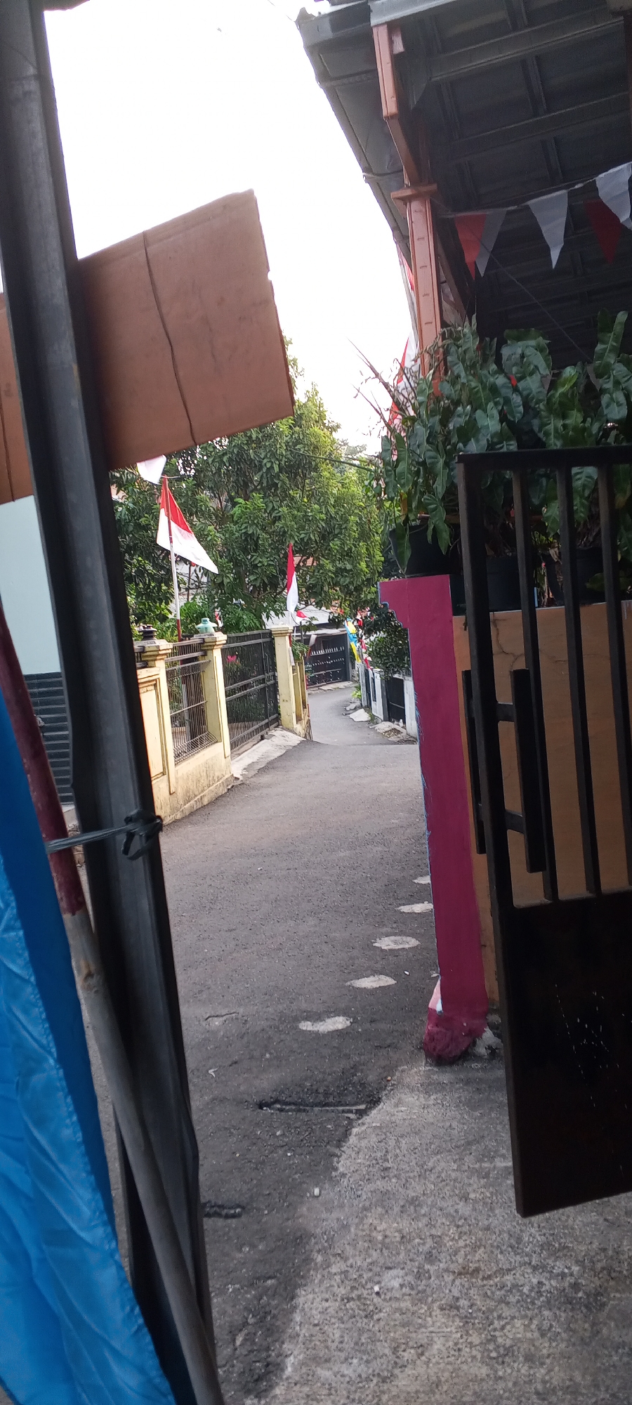 The image shows a street scene in Depok, Indonesia, likely a residential area, with Indonesian flags and decorations indicating a festive or national event. In the foreground, a vertical dark metal pole and a section of a blue tarp are visible on the left. The street is paved with asphalt and has some cracks and patches. On the right side of the street, a dark metal gate and a pink-painted pillar are present.
Further down the street, a yellow-colored fence with a black metal railing runs along the left side. Several Indonesian flags, characterized by their red and white horizontal stripes, are prominently displayed on poles behind the fence and further down the street. Trees with lush green foliage line the street, and some houses or buildings are visible in the background, some decorated with red and white triangular flags. The sky is overcast and bright. The overall impression is a typical street in a suburban or semi-urban Indonesian setting during a period of national celebration.