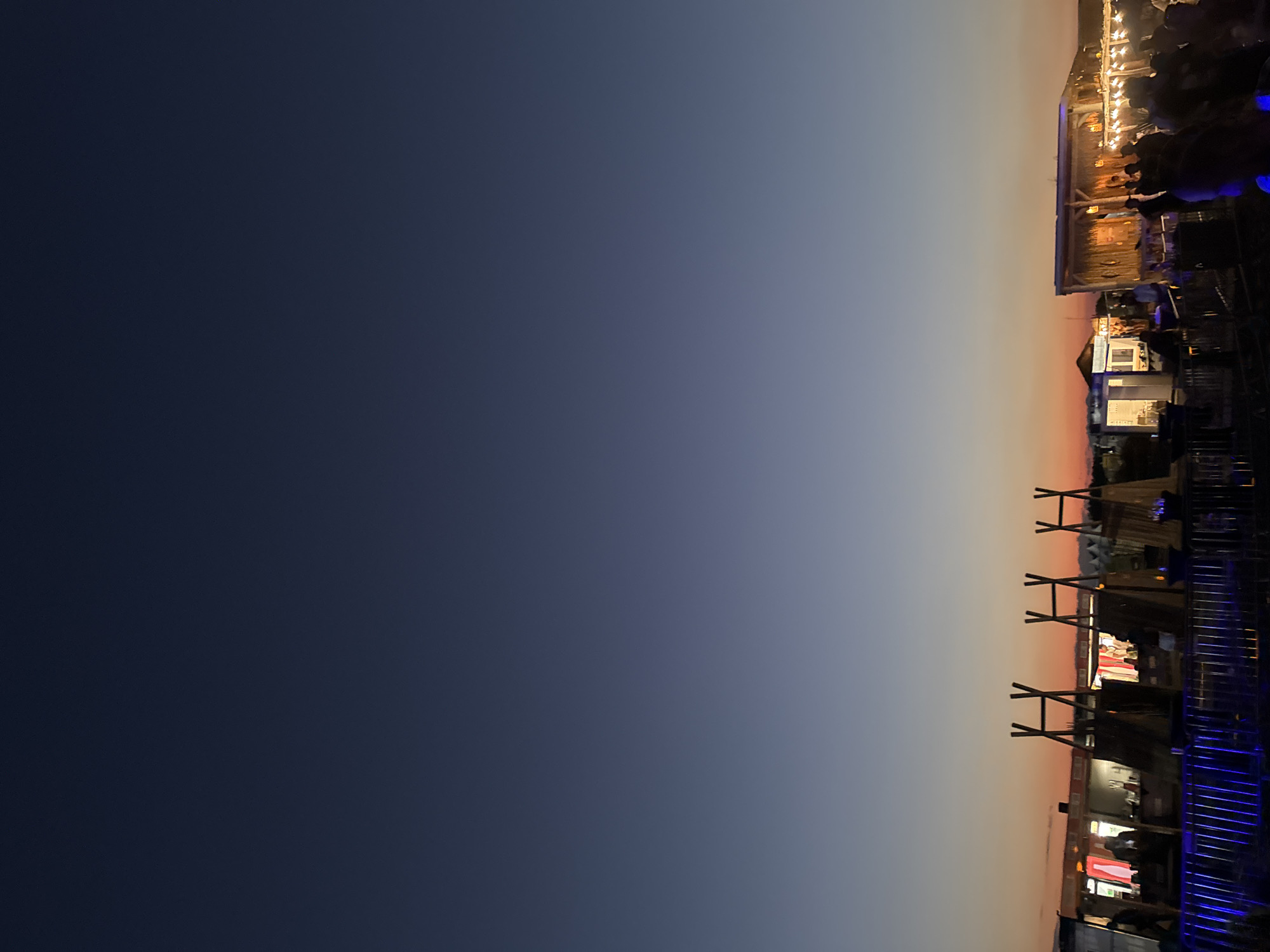 The image shows an outdoor scene at dusk with a gradient sky ranging from deep blue to orange. In the foreground, there is a railing with blue lights. Behind the railing, there are several structures that appear to be stalls or food stands, some with thatched roofs. Some of these structures have wooden frames that resemble teepees. People are visible in the background, gathered around the stalls, some of whom appear to be customers or vendors. The overall setting suggests a market, festival, or outdoor gathering. The location is identified as Donji Štoj, Montenegro.