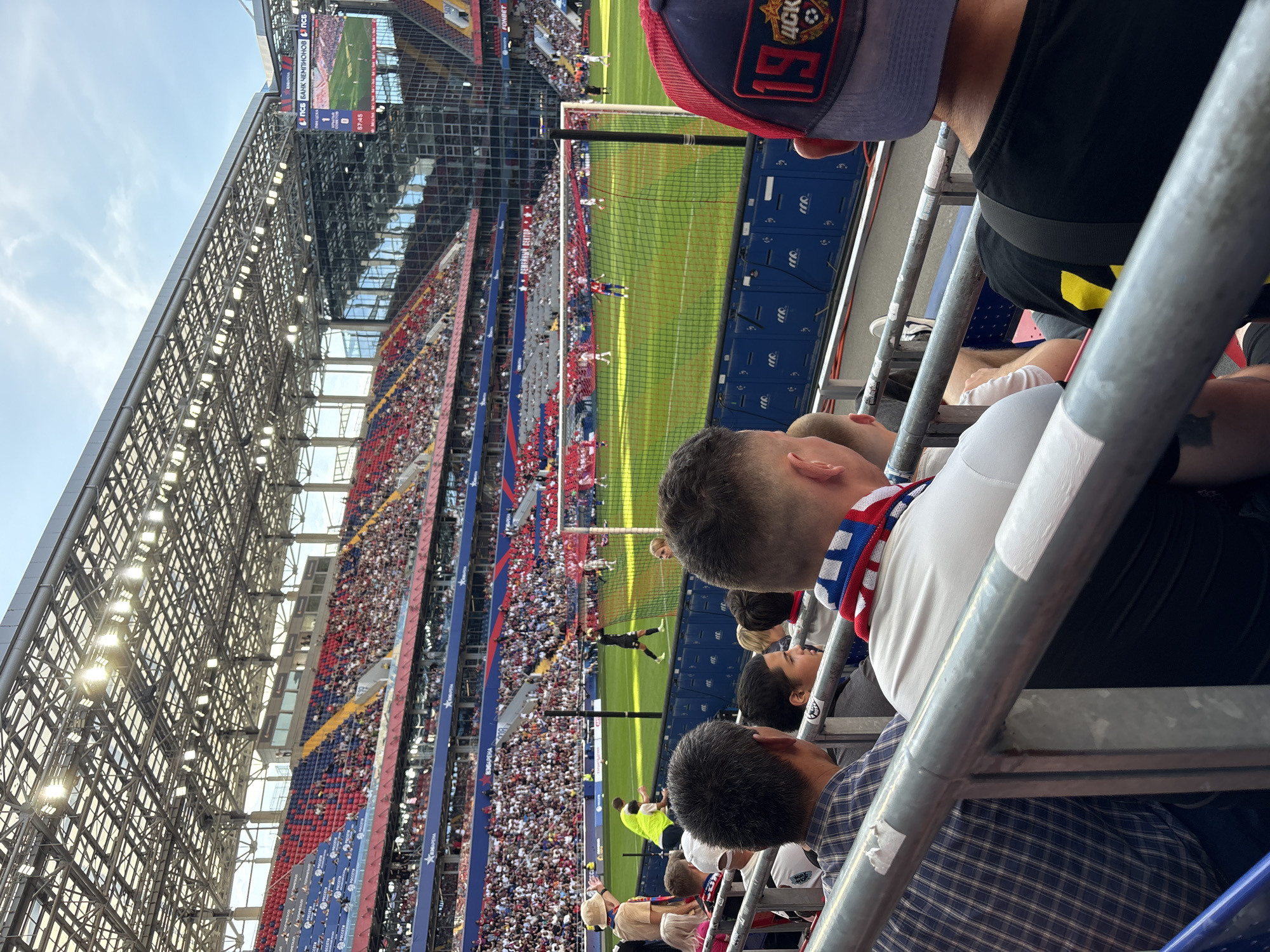 In the city of Khimki, Russia, this image captures a football match in progress within a modern stadium. The foreground features spectators seated in tiered rows, partially obscured by metal railings. A man in the immediate foreground wears a dark baseball cap with a red brim and a logo featuring the number '19' and Cyrillic lettering. To his left, another spectator is visible from the back, wearing a white shirt and a red, white, and blue scarf. Further to the left, a man in a blue plaid shirt is seated.
The middle ground of the image reveals the football field bathed in sunlight, with players in white and dark uniforms actively engaged in the game. A goal net is visible on the right side of the field. Spectators fill the surrounding grandstands, which are colored red and blue, indicating team affiliations.
The background shows the stadium's infrastructure, including a complex metal roof structure supporting numerous bright lights. A large video screen is mounted above the stands on the right, displaying a live feed of the match. The overall environment is that of a professional sporting event, with a clear sky overhead.