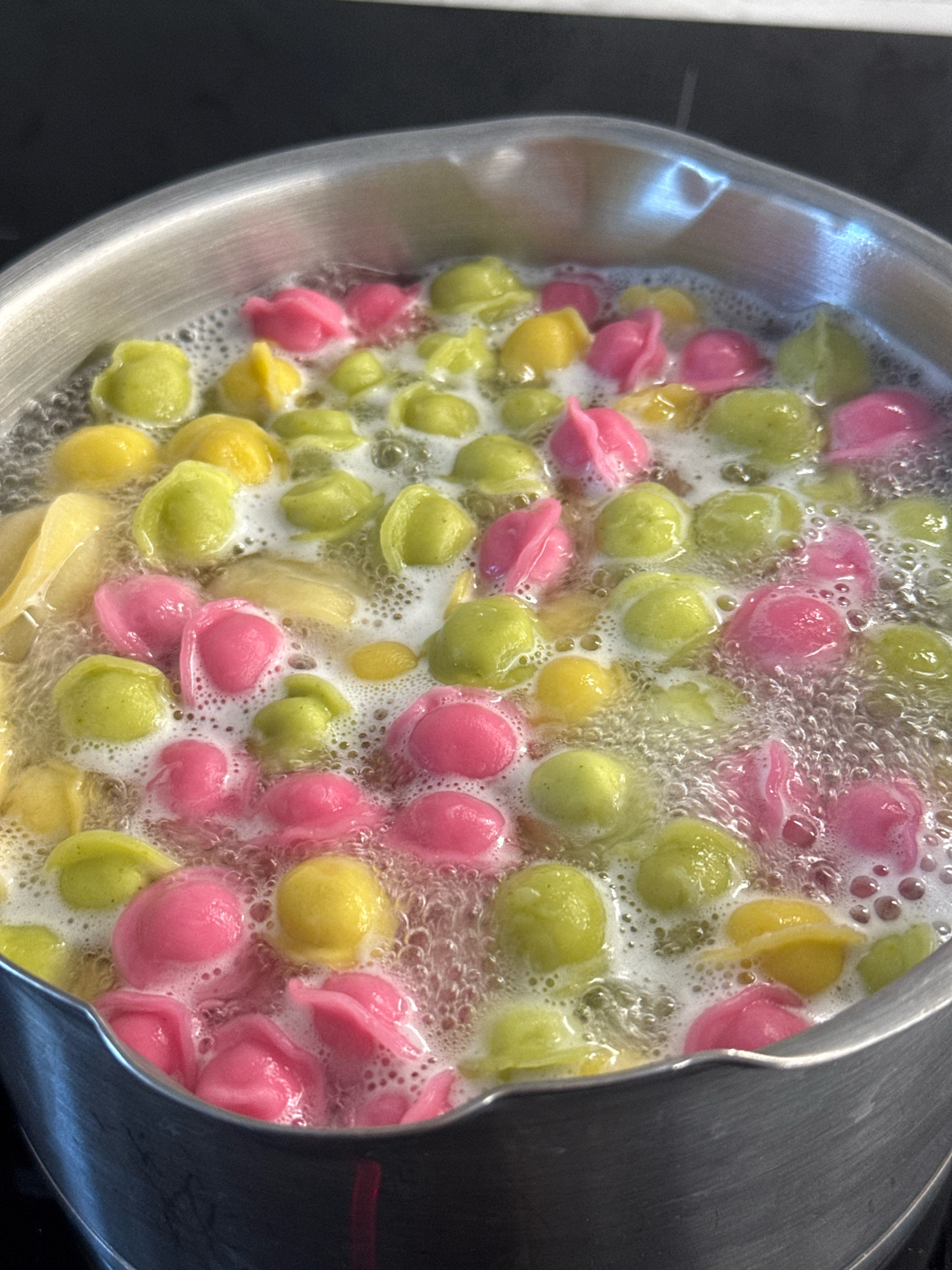 A high-angle, close-up shot shows colorful dumplings with a petal-like shape, colored pink, yellow, and green, boiling in a stainless steel pot. The dumplings are partially submerged in bubbly water, with some floating to the surface. The pot is filled with a significant amount of water, indicating the dumplings are being cooked. The background is dark and out of focus, suggesting the scene is indoors, possibly in a kitchen. There is a reflection of bright light on the rim of the pot. No people are visible. The image does not contain any specific visual information that could be used to determine its location in Lyubertsy, Russia.
