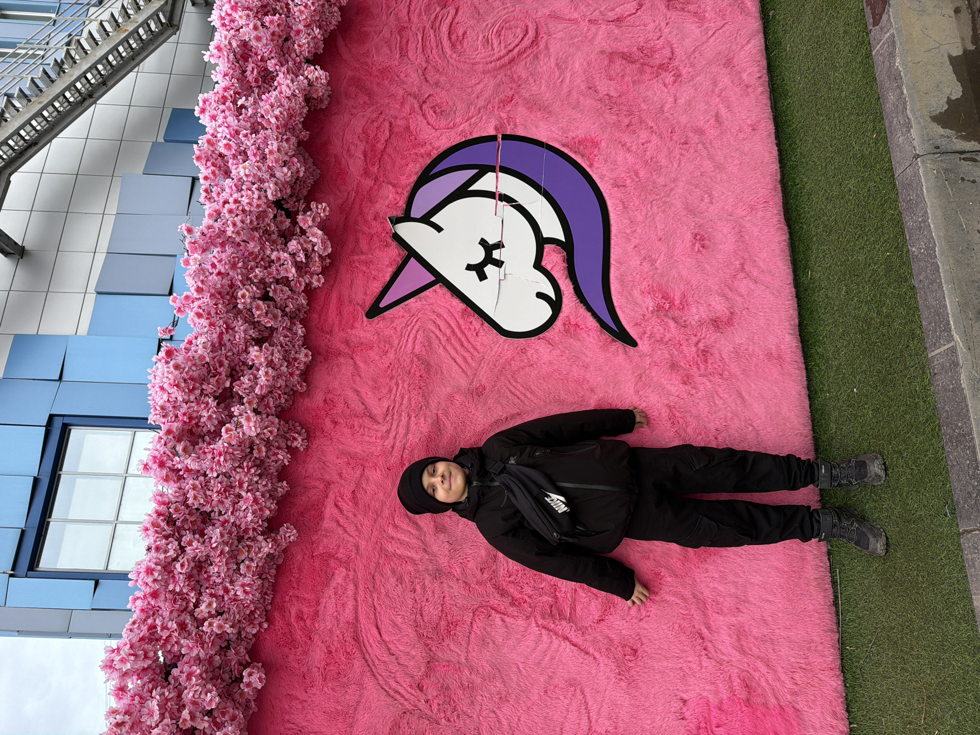 A photograph, taken outdoors in Votkinsk, Russia, depicts a young person lying on a large, pink, shaggy rug with a cartoon unicorn head on it. The person is wearing a black beanie, jacket, pants, and shoes, as well as a black Nike waist pack. To the left of the pink rug is a building facade with pink floral decorations and a metal fire escape. To the right of the rug is a patch of artificial green turf, followed by a sidewalk. The unicorn head on the rug is white with a purple mane and horn, outlined in black. A thin, red string stretches upward from the unicorn's head.