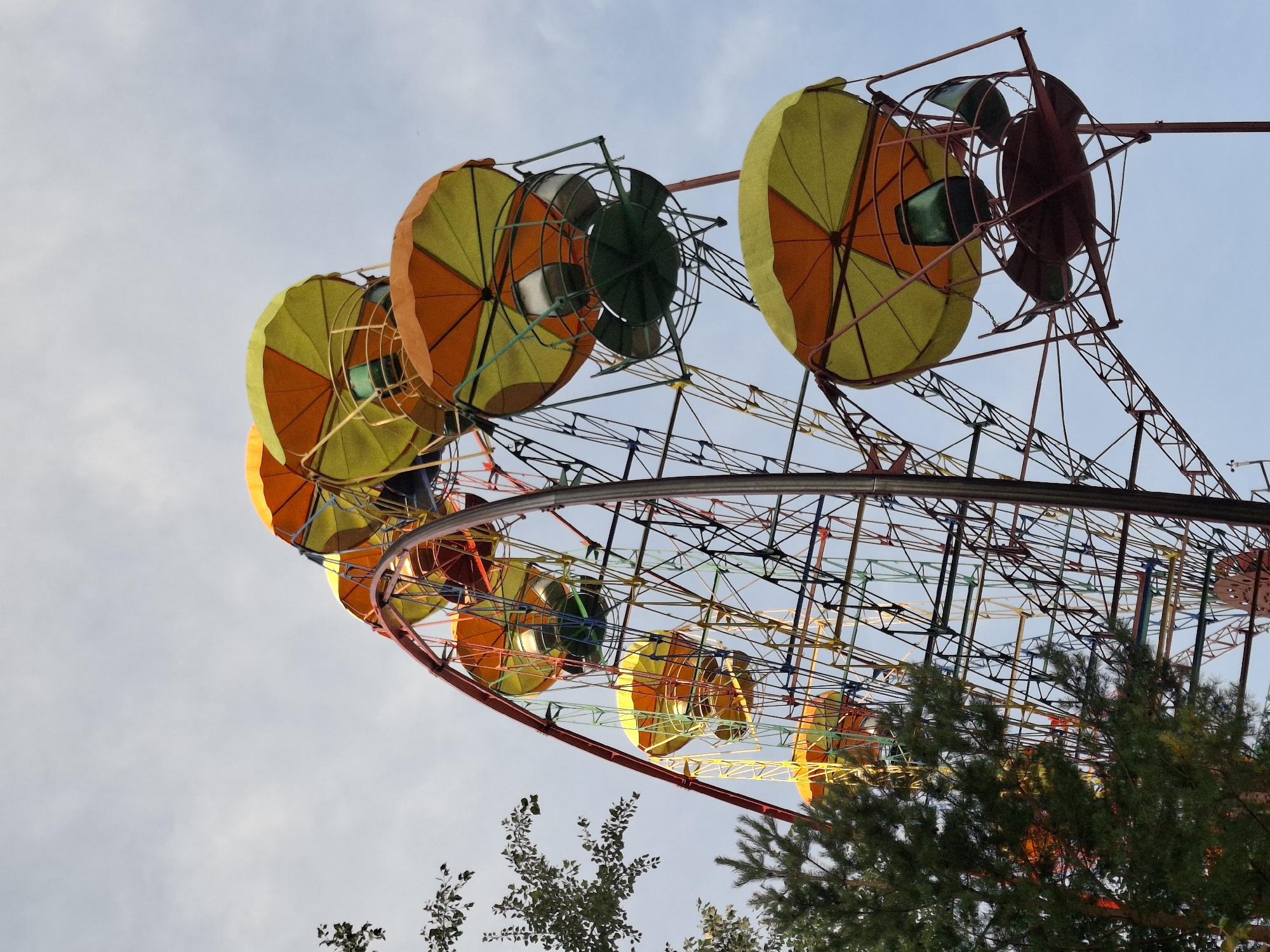 The image depicts a Ferris wheel, a type of amusement ride consisting of a rotating upright wheel with multiple passenger-carrying components attached to the rim in such a way that as the wheel turns, they are kept upright, usually by gravity. This Ferris wheel is colorful, with yellow and orange canopies over its gondolas. It is located in the city of Chaikovsky, Russia. The sky is partially cloudy, and trees are visible in the foreground, suggesting an outdoor setting in a park or amusement area. There are no people visible in the image, and no actions are taking place. The Ferris wheel appears to be stationary.