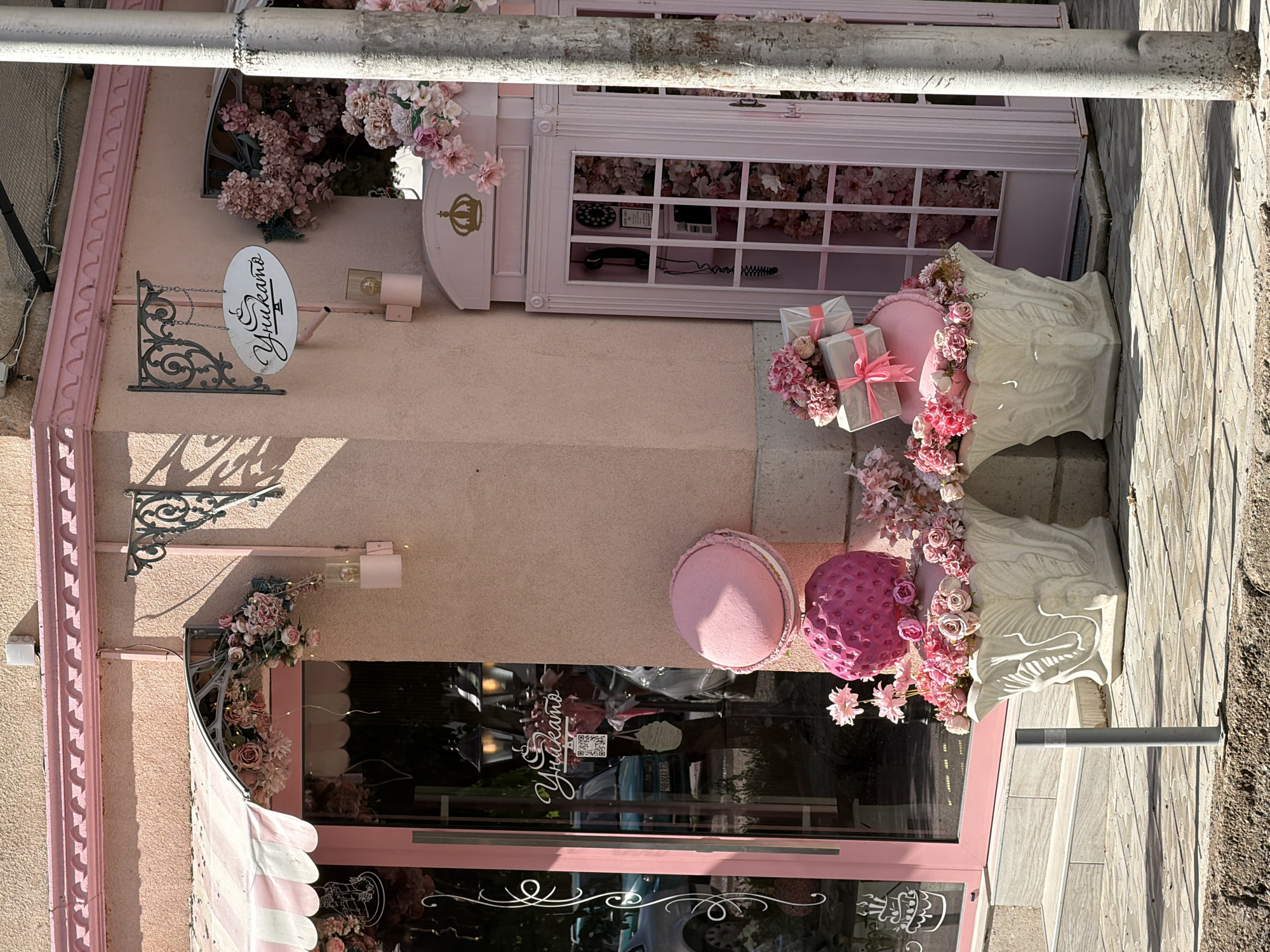 This image shows the exterior of a business, likely a pastry shop or bakery, named "Yukaryano," located in Pleven, Bulgaria. The building is painted in various shades of pink and decorated with flowers.
Architectural features include a pink facade with decorative molding, a door with a striped awning, and a glass storefront. A sign with the name "Yukaryano" is mounted on the building. There is a decorative telephone box, also painted pink.
The sidewalk in front of the building is visible.
The image is taken during daylight hours.