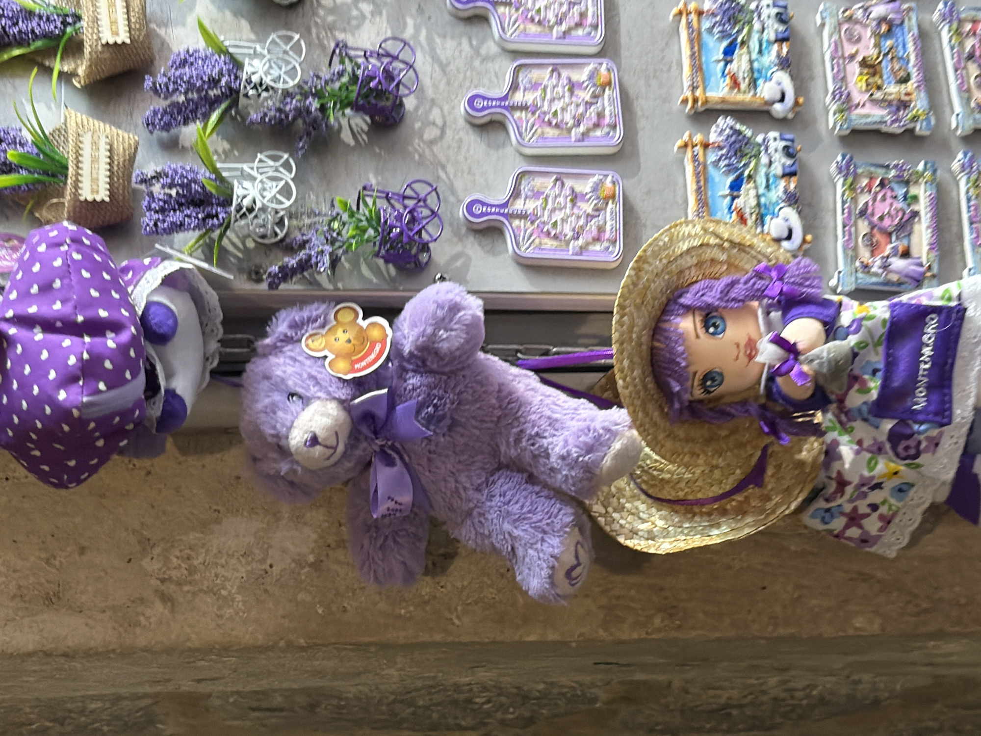 The image displays a close-up view of various novelty items, likely souvenirs, affixed to a textured wall. In the upper left quadrant, a purple stuffed bear with a ribbon around its neck is prominently featured. Below the bear, a doll wearing a straw hat and a floral dress is visible. To the right of these figures, several items with lavender motifs are arranged, including small baskets with artificial lavender sprigs, decorative magnets resembling cutting boards with lavender scenes, and rectangular magnets depicting coastal landscapes with lavender fields and traditional architecture. The overall color palette is dominated by shades of purple and the natural tones of the wall and straw hat. While the specific location within Budva, Montenegro is not explicitly shown, the presence of items with "MONTENEGRO" labeling suggests a tourist-oriented context. The items are static, and no actions or interactions are occurring. The environment appears to be an indoor setting, possibly a shop or market stall.